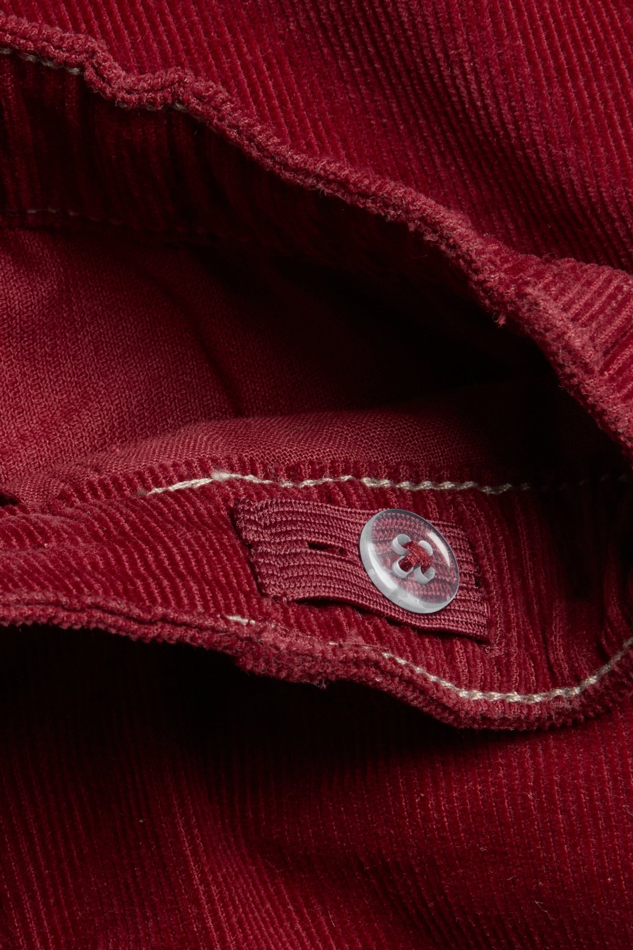 Close-up of red corduroy trousers showing a clear button and elastic tab on the waistband.