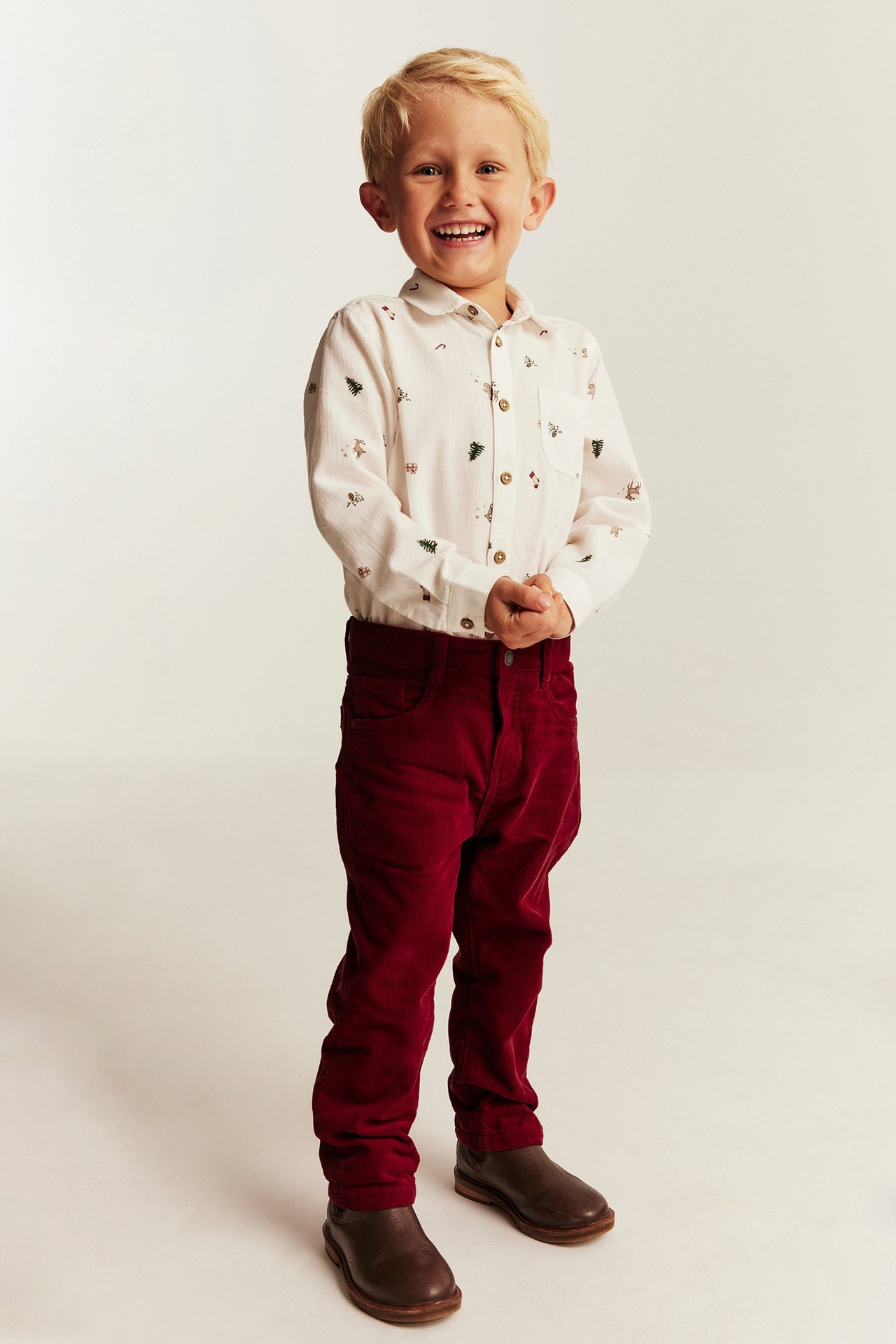 Front view of a kid in a white festive print long-sleeve shirt, red corduroy pants, brown shoes.