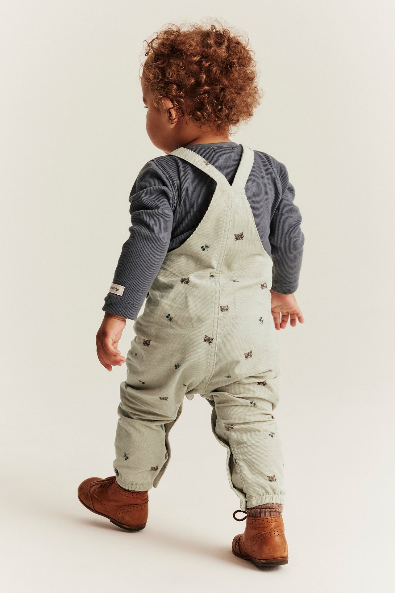 Rückenansicht eines Babys in beiger gemusterter Cord-Latzhose über einem dunkelgrauen Langarmshirt und braunen Schuhen.