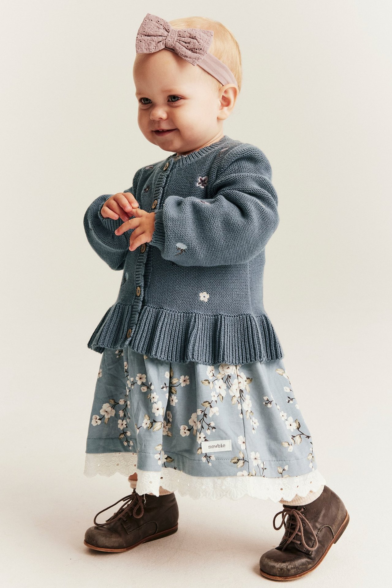 Vorderansicht: Baby in blauer bestickter Strickjacke, geblümtem Kleid, rosa Schleife und braunen Stiefeln.
