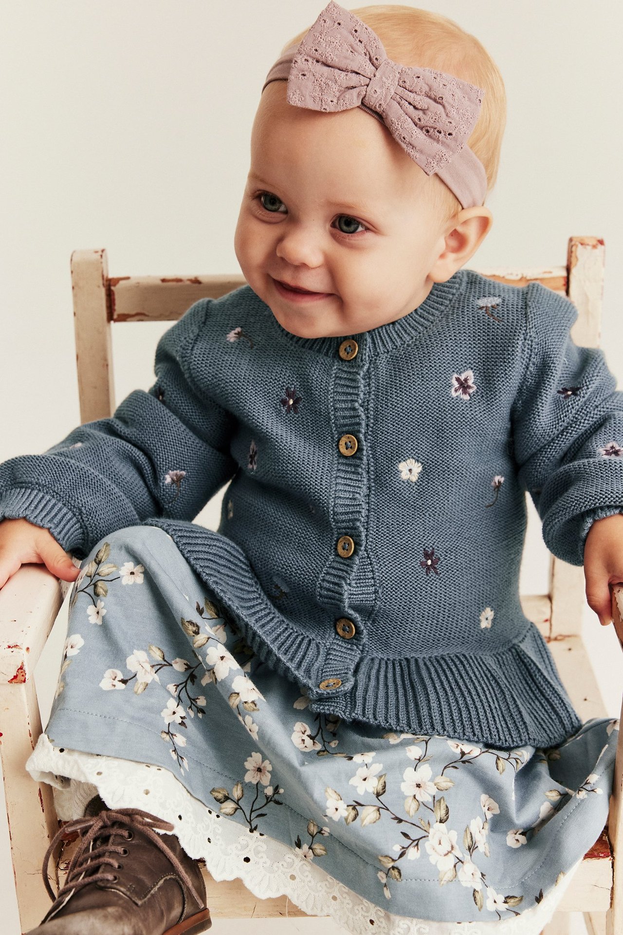 Vorderansicht eines Babys in blauem gestricktem besticktem Cardigan, Blumenkleid und rosa Schleifen-Stirnband.