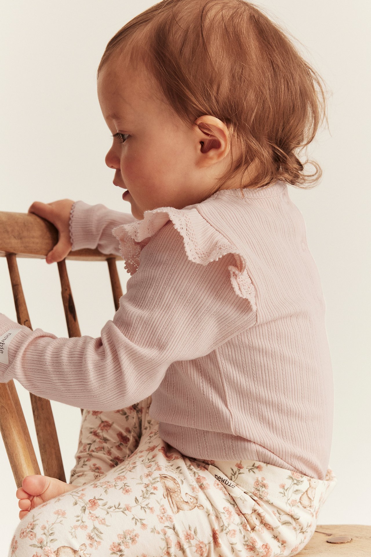 Side view of baby wearing a light pink ribbed long-sleeved body with lace-edged ruffles and floral leggings.