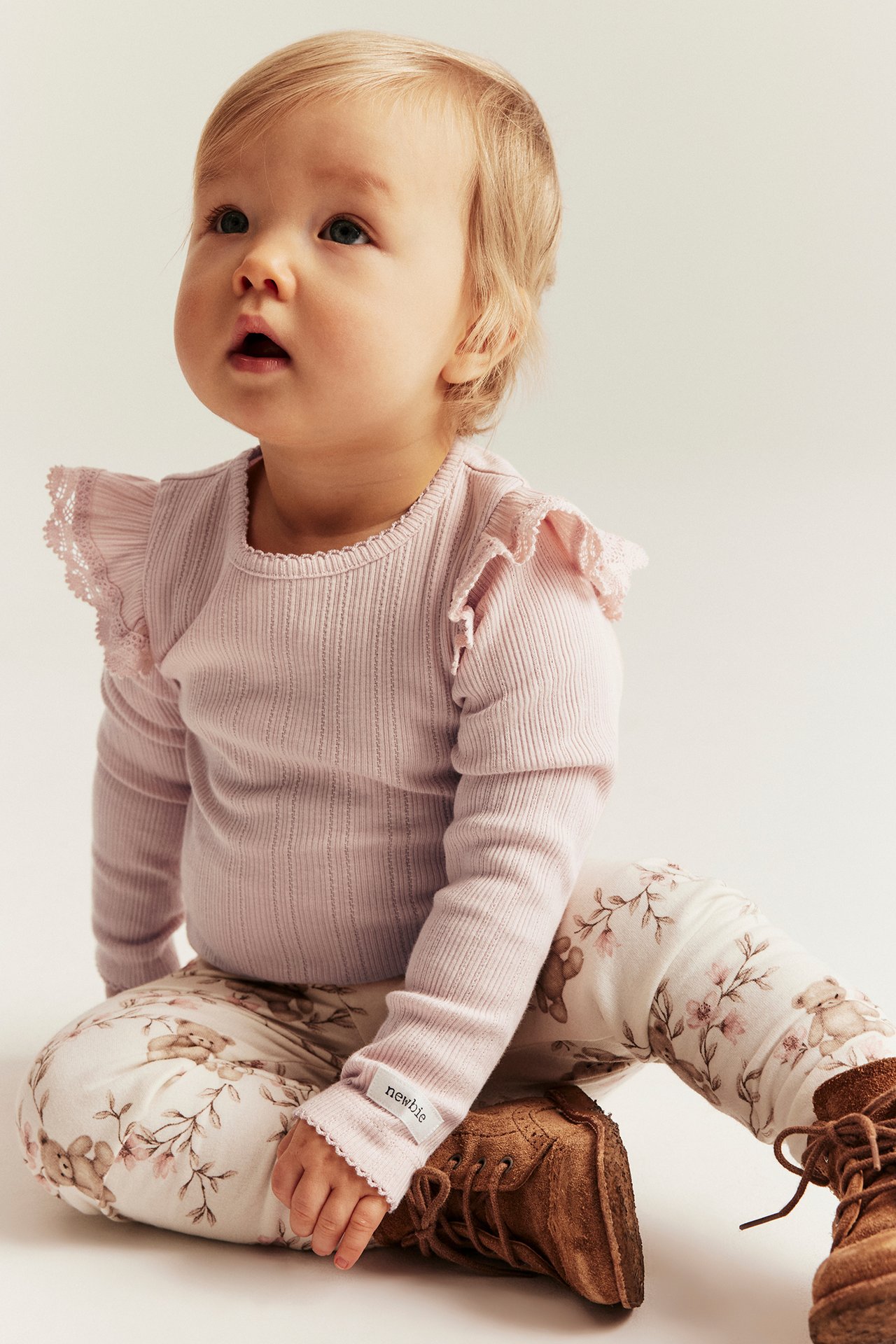 Front view of baby in a light pink ribbed bodysuit with ruffled shoulders, patterned leggings, and brown boots.