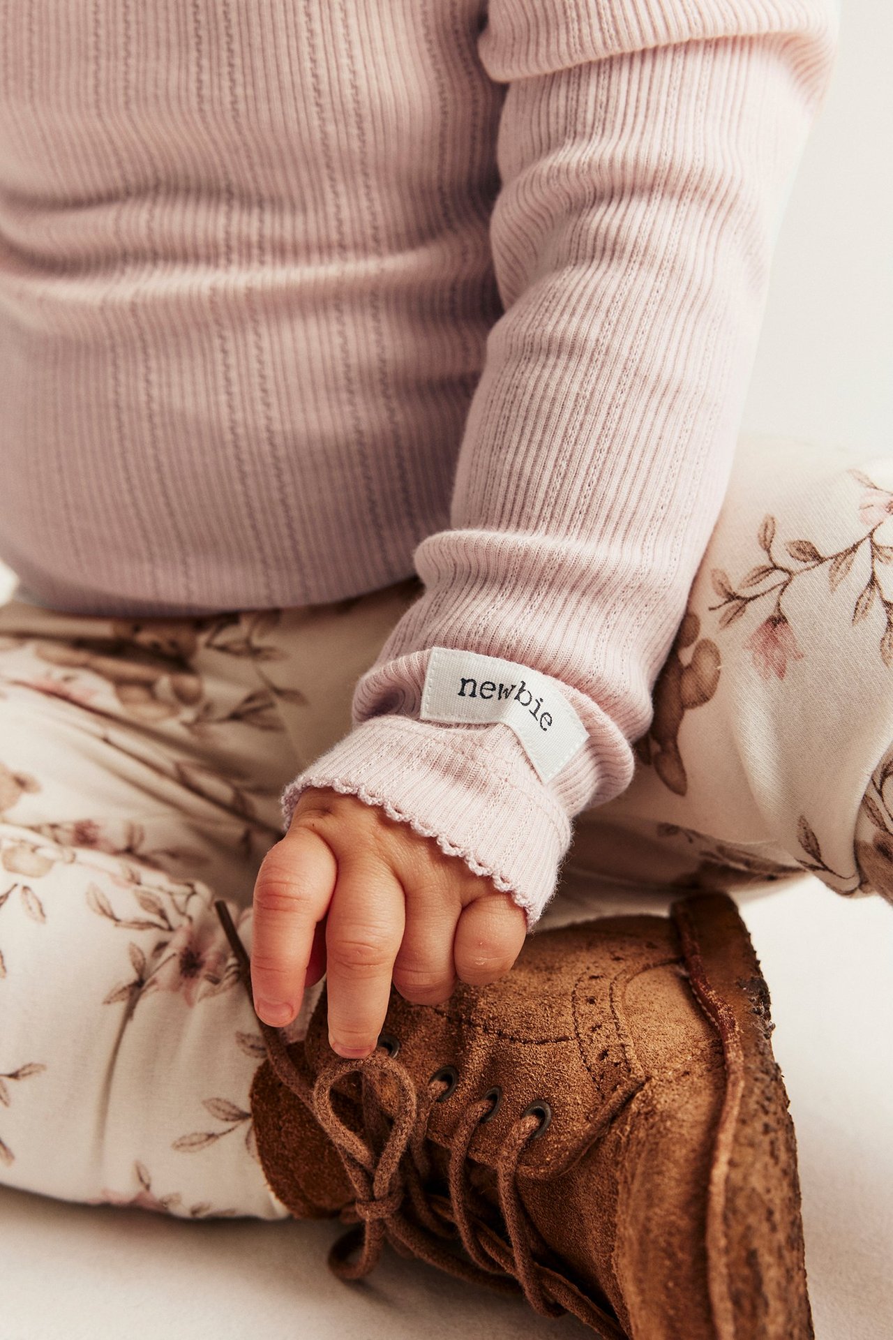Close-up of baby's light pink ribbed bodysuit with scalloped cuffs, floral pants, and brown laced boot.