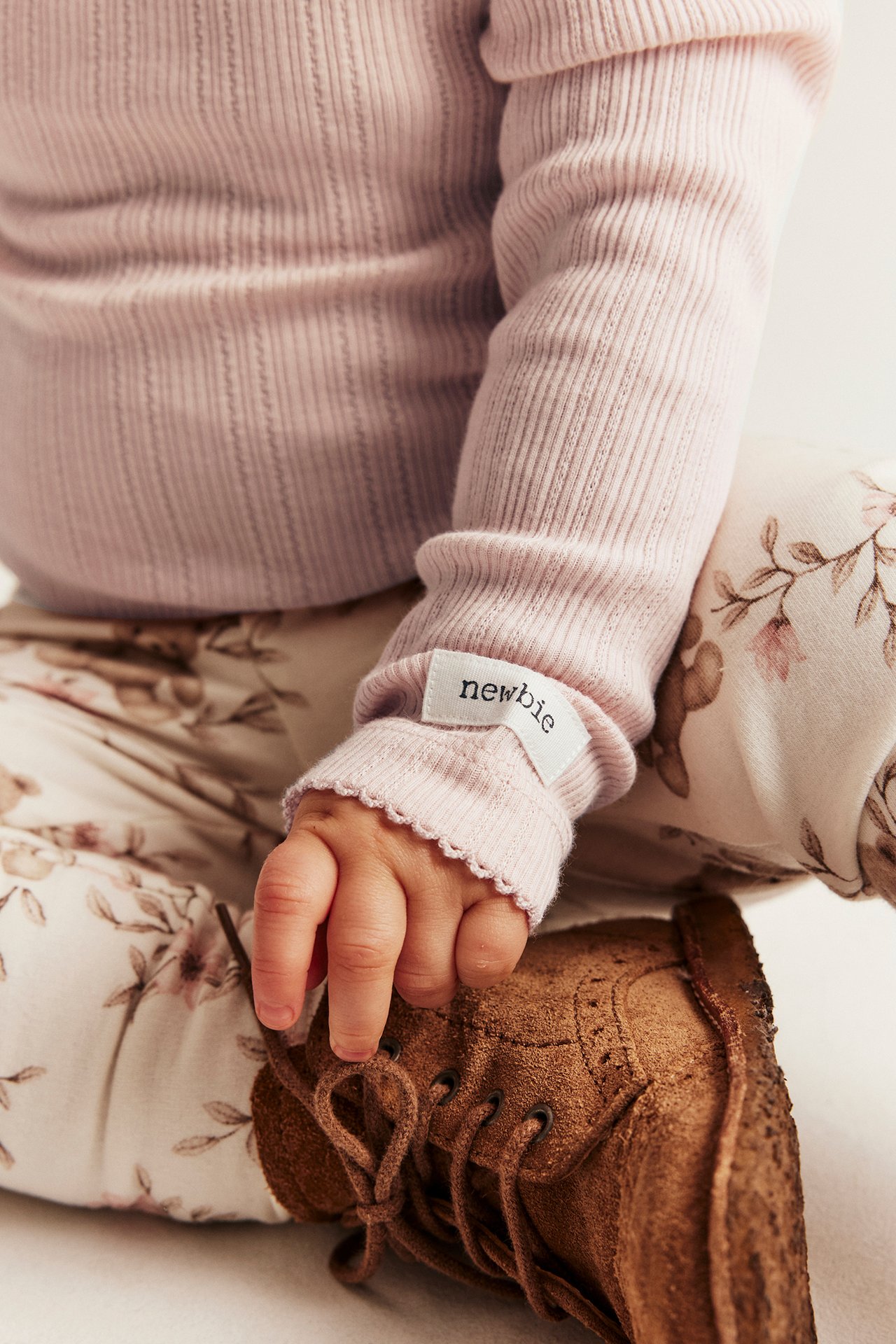 Nahaufnahme von Baby in hellrosa Ripp-Body mit gewelltem Ärmelbündchen, geblümter Hose und braunem Schnürstiefel.
