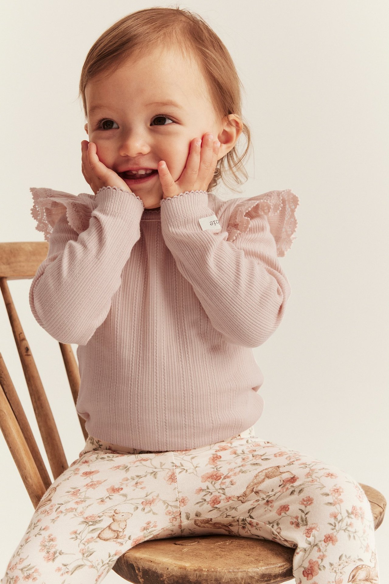 Front view of a baby wearing a light pink ribbed body with lace-edged shoulder ruffles and patterned leggings.