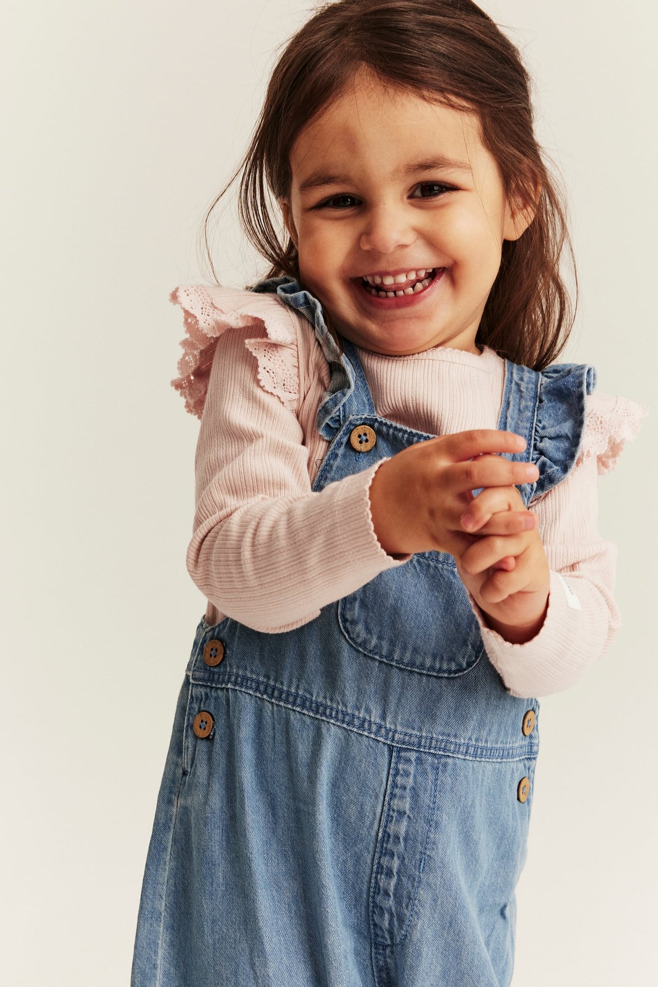Vorderansicht, lachendes Kind in hellrosa geripptem Rüschen-Body und hellblauer Jeanslatzhose, klatscht Hände.