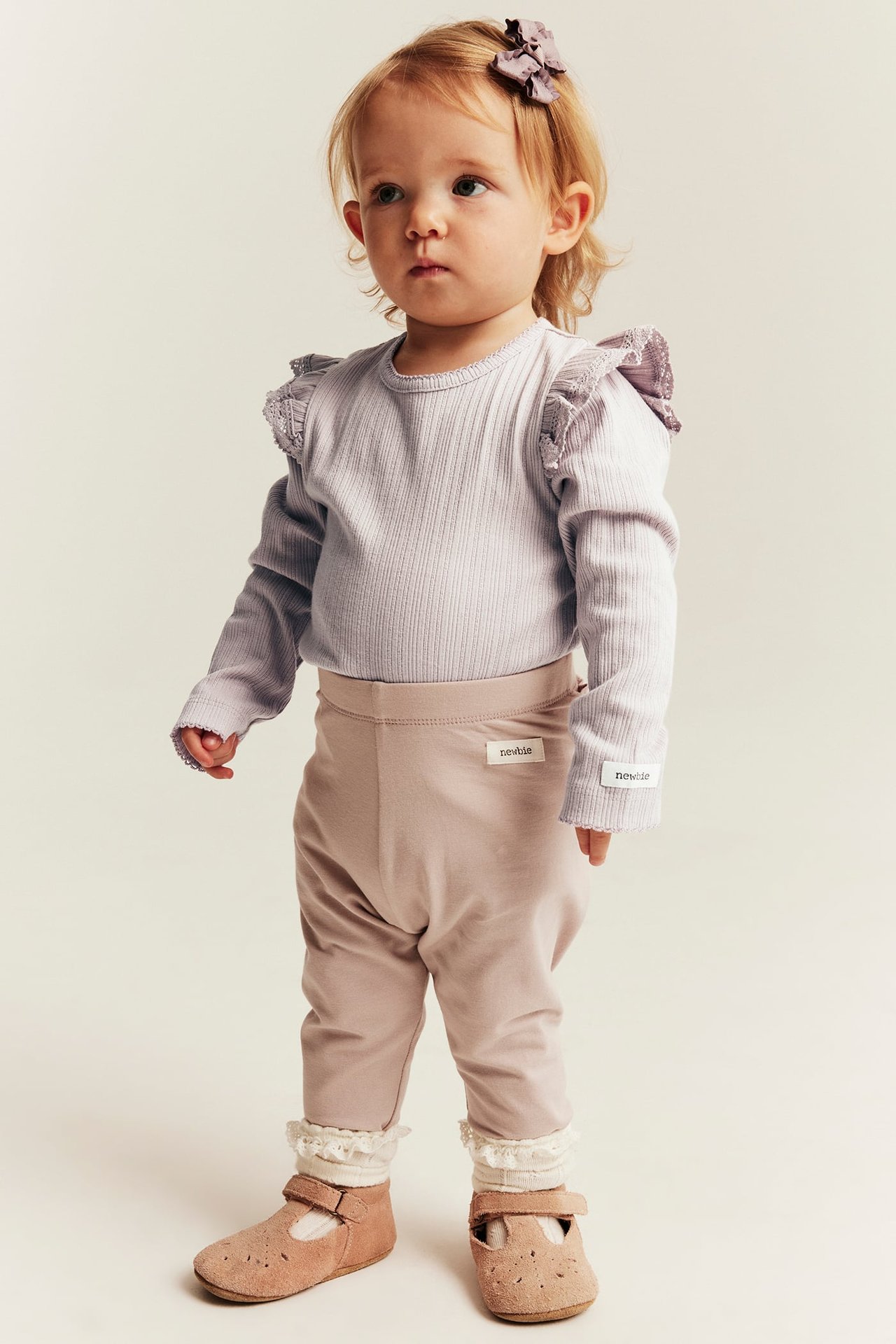 Baby in light purple ribbed ruffle top, light brown leggings, cream socks, brown shoes, purple bow.