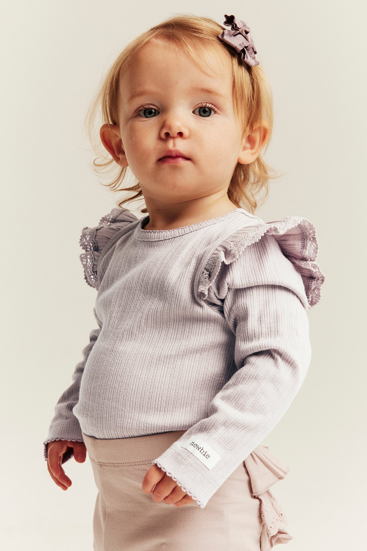 Front view: baby in light purple ribbed top with ruffled shoulders, beige pants, purple hair bow.