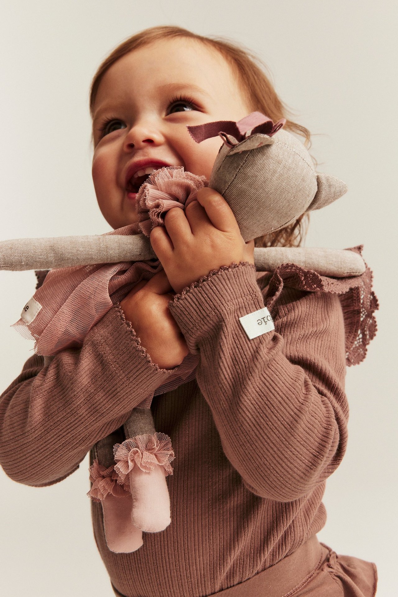 Nahaufnahme eines Babys in einem dunkelrosa gerippten Body mit Spitzenrüschen, hält eine Stoffpuppe.