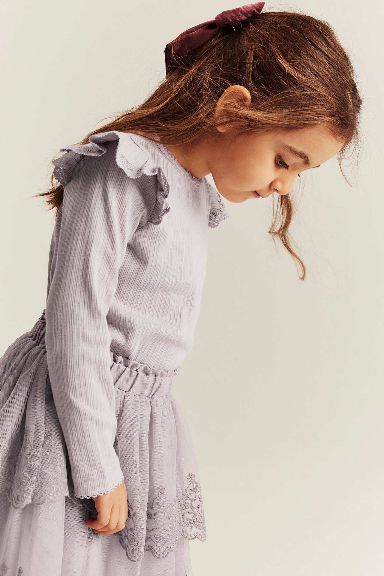 Side view of a kid wearing a light lilac ribbed long-sleeved top with lace-edged ruffles and a purple tulle skirt.