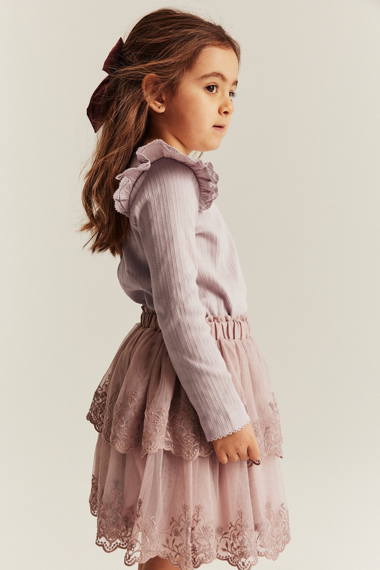 Seitenansicht Kind in helllila geripptem Oberteil mit Schulterrüschen, pinkem Stufenrock und dunkelroter Schleife.