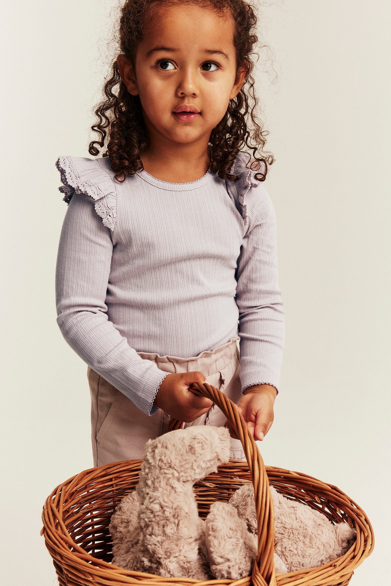 Front view: Kid in a lilac ribbed long-sleeved top with ruffles, light beige pants, holding a toy basket.