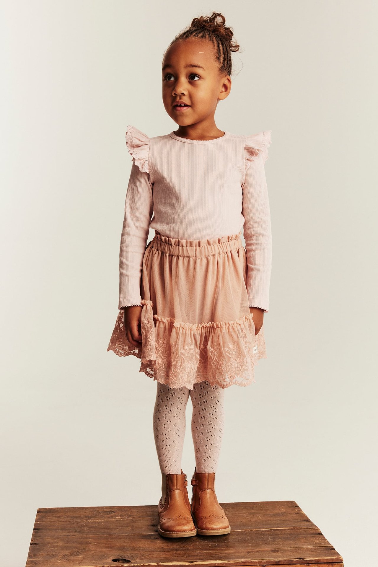 Front view of kid in pink tiered lace skirt, pink ribbed ruffle top, white patterned tights, brown boots.