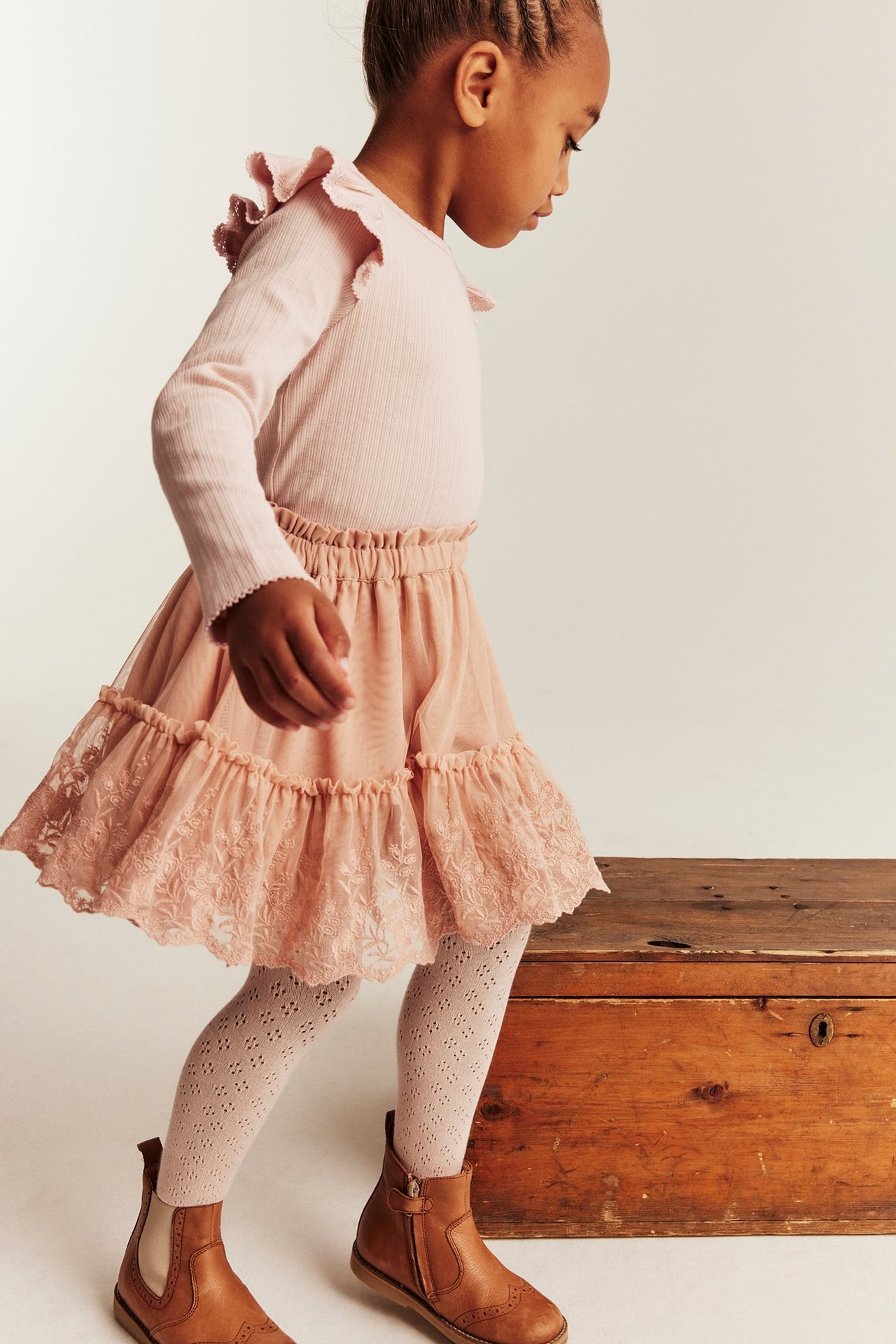 Kid wears light pink tiered lace skirt, pink ribbed top, patterned tights, brown boots.