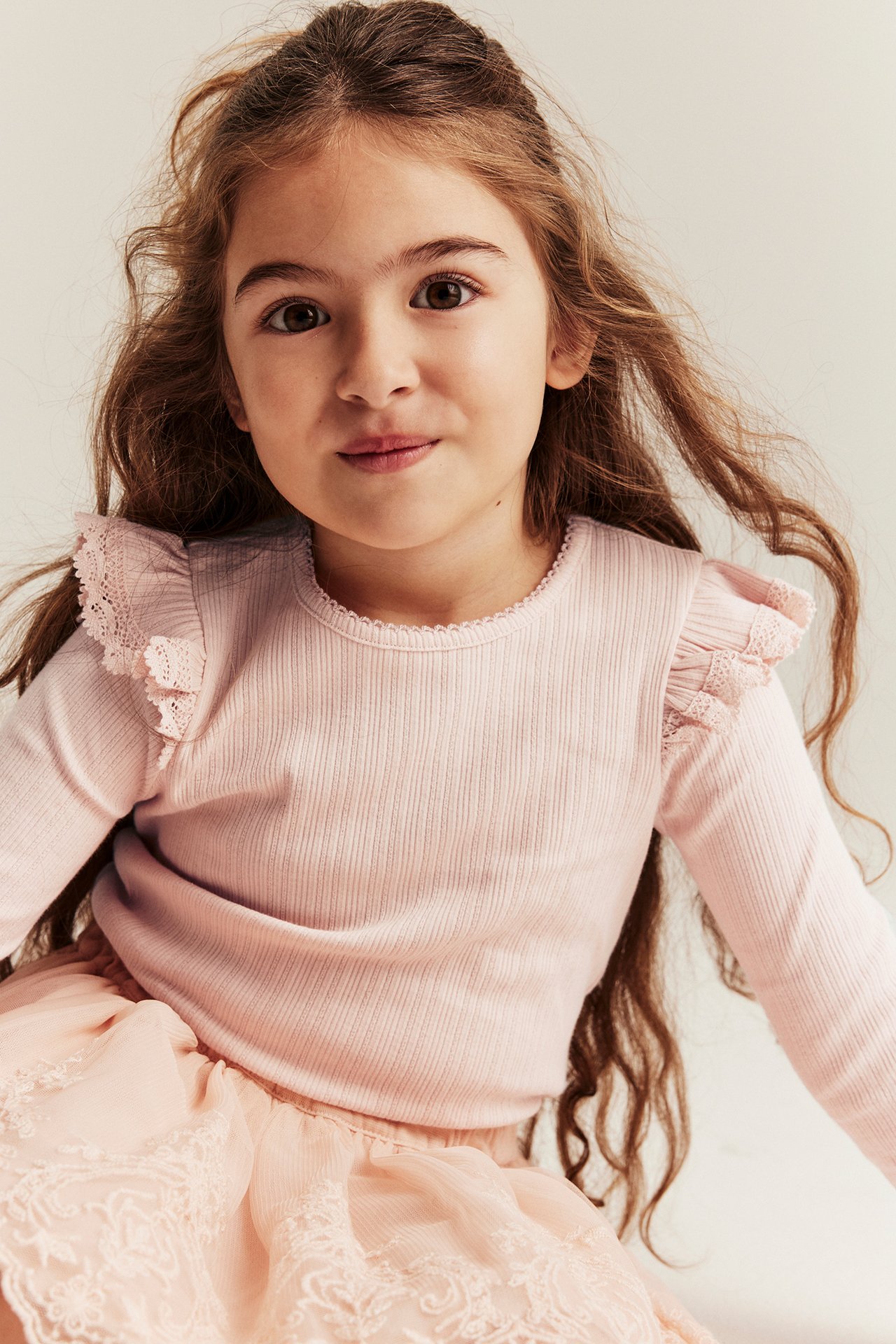 Close-up: Kid in light pink ribbed long-sleeve top with lace ruffles and a pink lace skirt.
