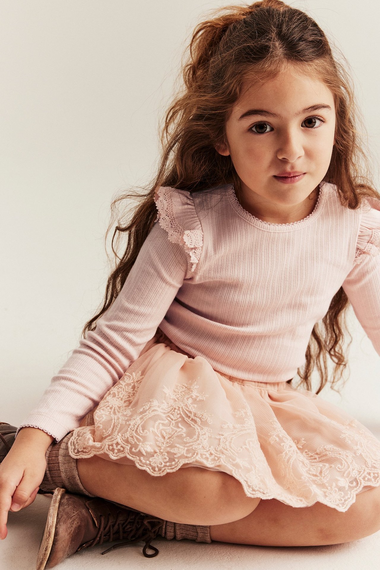 Vorderansicht: Mädchen trägt hellrosa geripptes Top mit Spitzenrüschen, bestickten Rock und braune Schuhe.