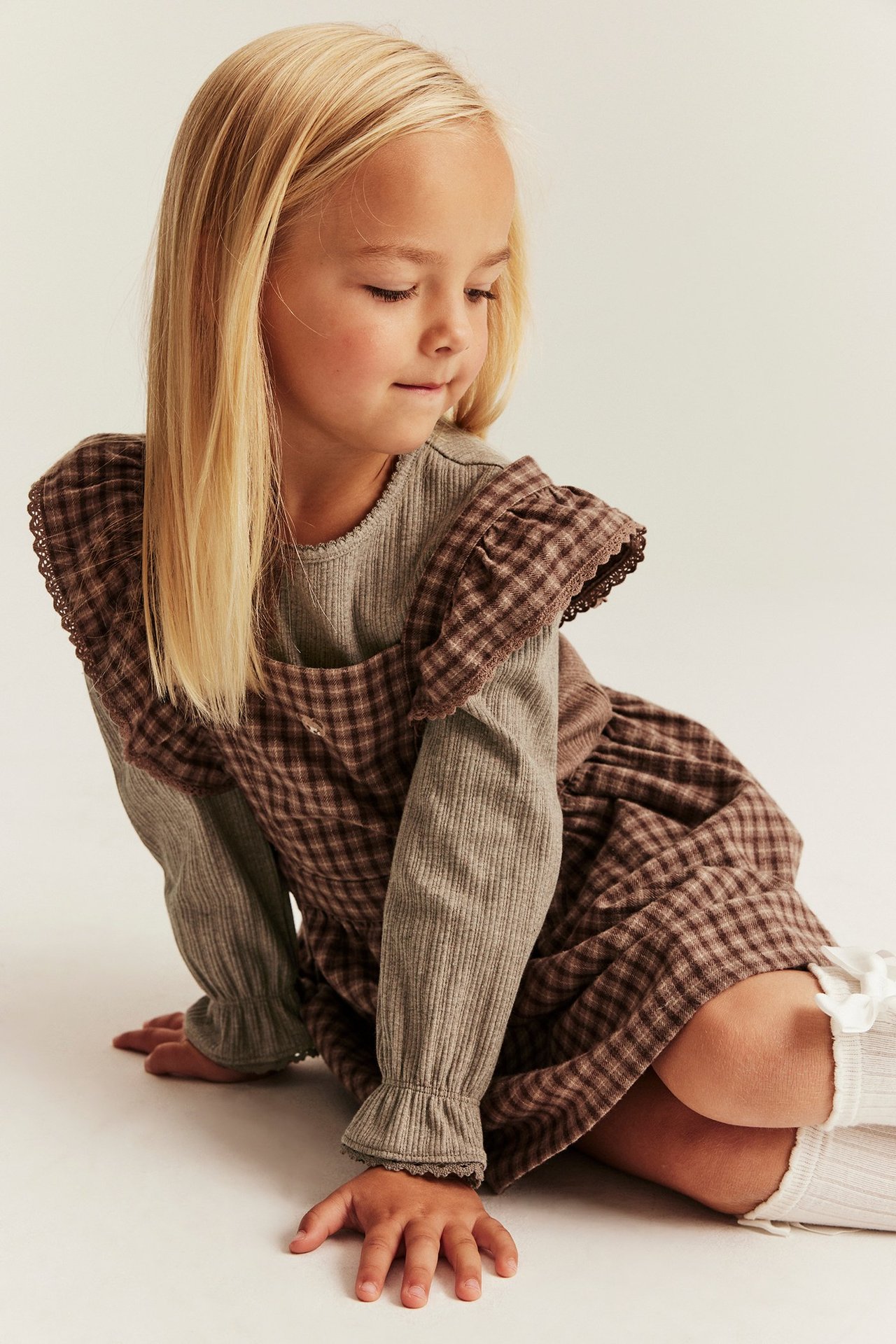 Vorderansicht: Kind in hellbraunem geripptem Langarmshirt mit Spitzenbündchen, unter kariertem Kleid.