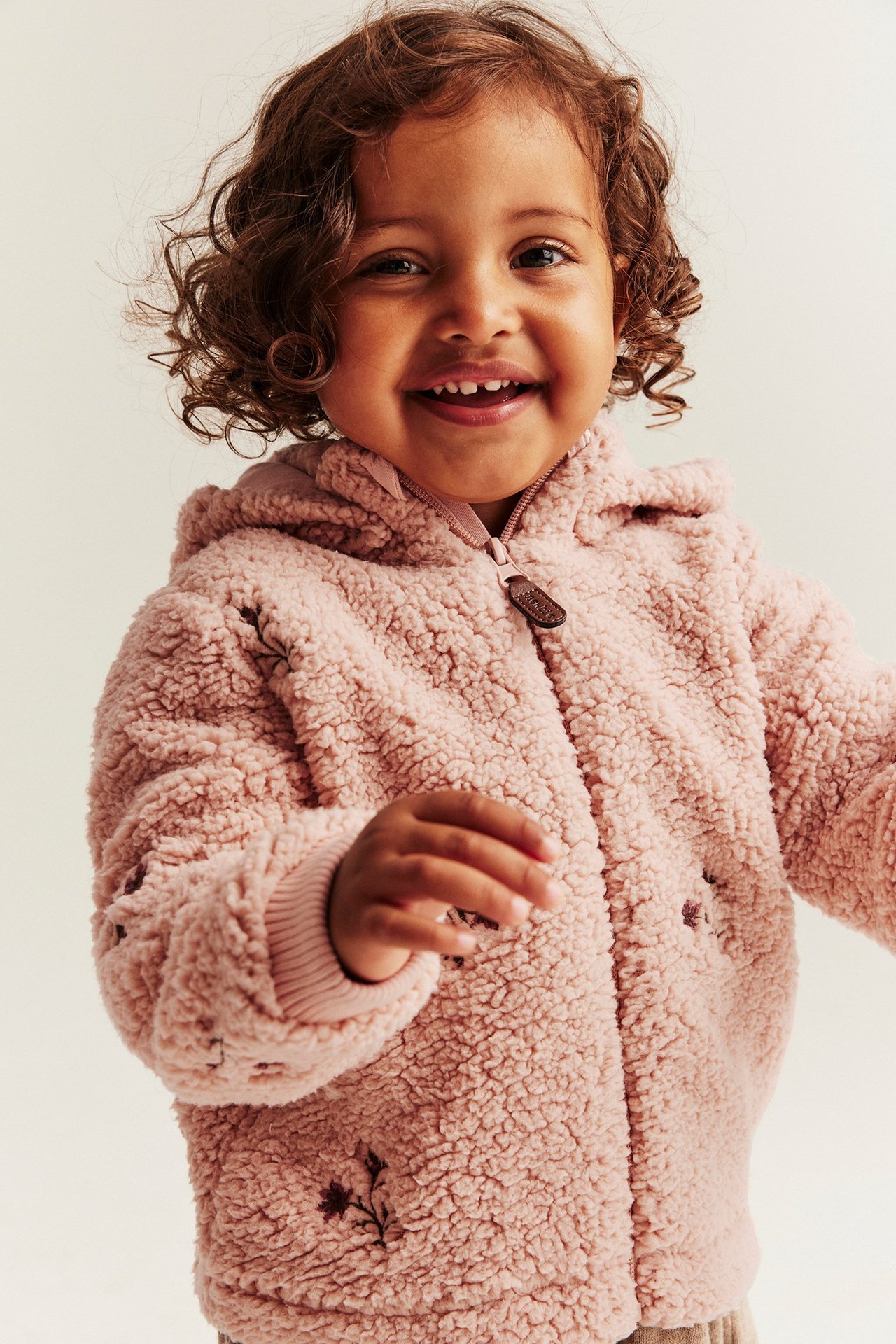 Front view of a smiling kid wearing a pink pile jacket with floral embroidery and a zipper.