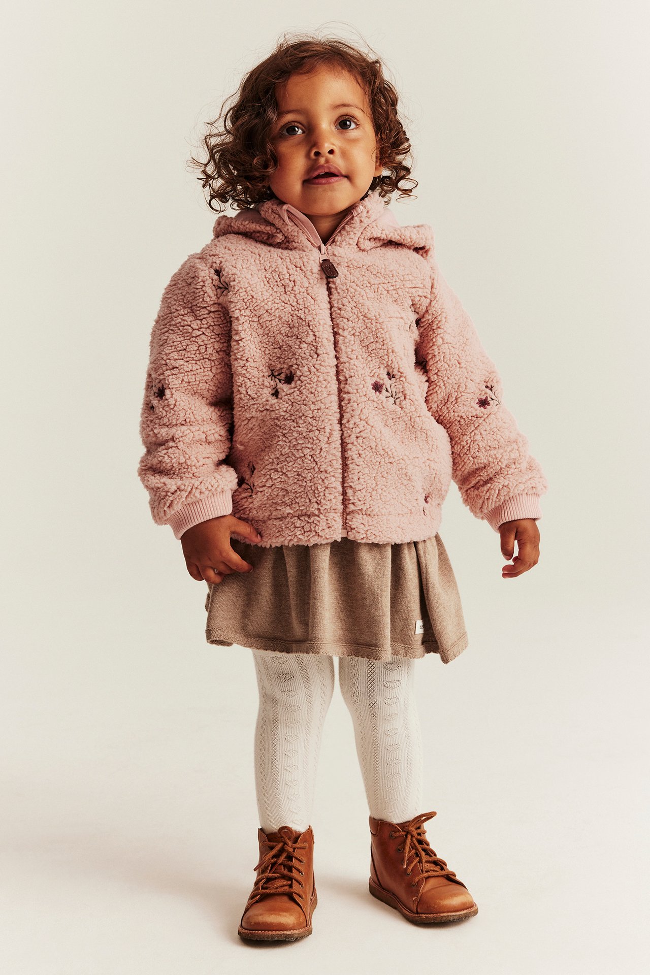 Front view: kid in pink hooded teddy jacket with flowers, beige skirt, white tights, brown boots.