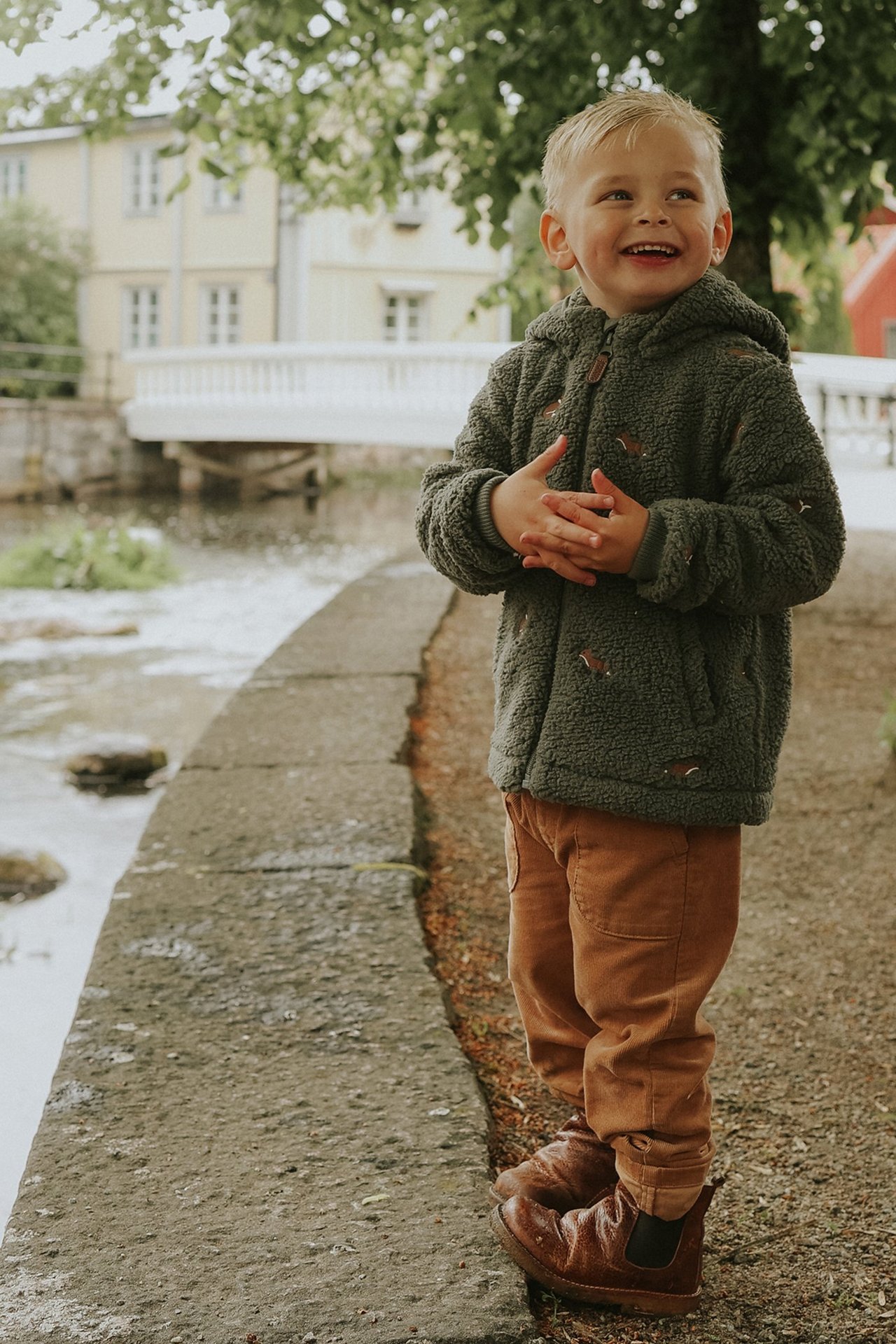 Vorderansicht eines lächelnden Kindes in dunkelgrüner Fleecejacke mit Fuchsstickerei, brauner Cordhose und braunen Stiefeln.
