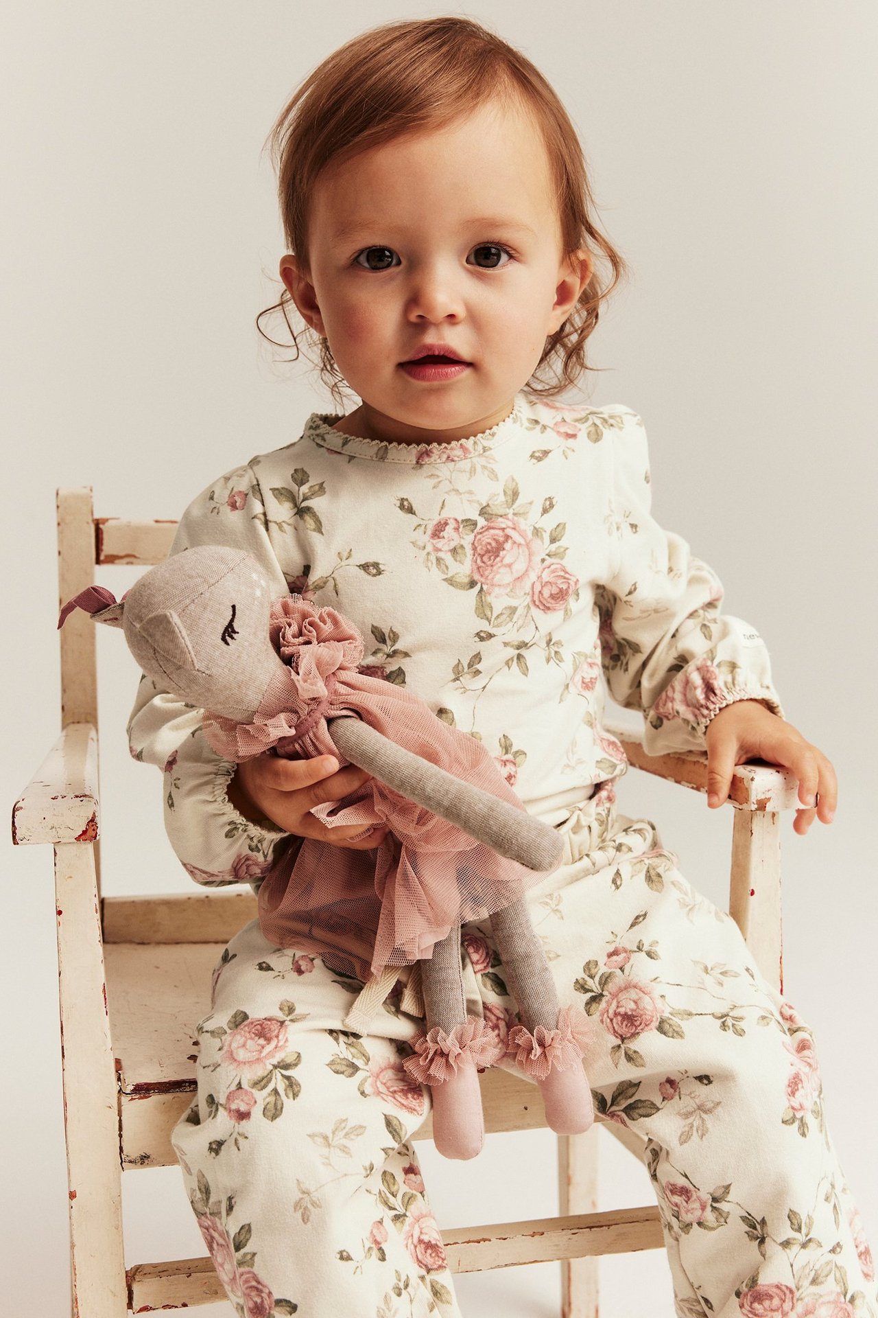 Vorderansicht eines Babys in hellbeigem Blumen-Puffärmel-Body und passender Hose, das eine rosa Puppe hält.