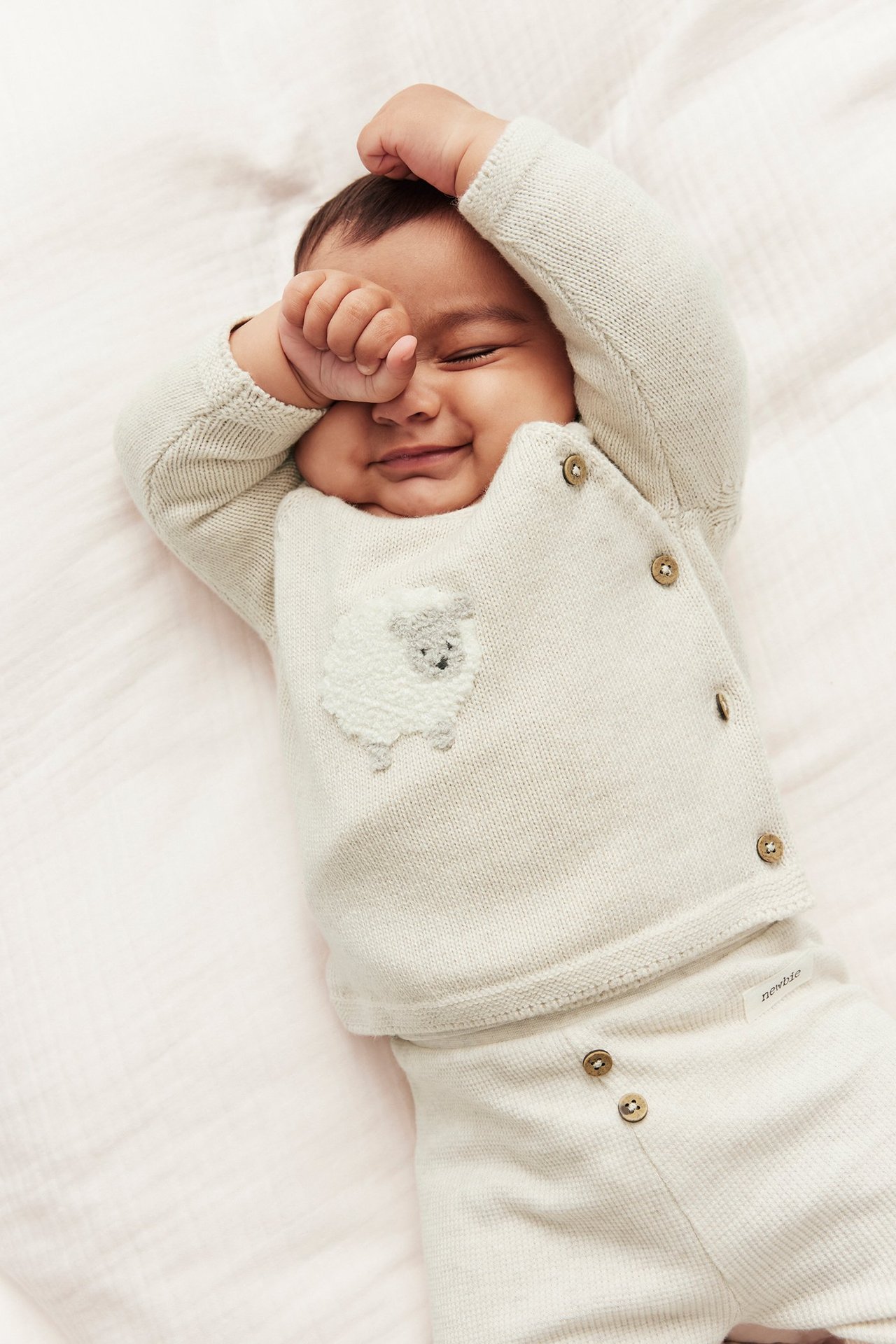 Draufsicht: Baby in beiger melierter gestrickter Baumwoll-Wickeljacke mit Schaf-Applikation und passender Hose.