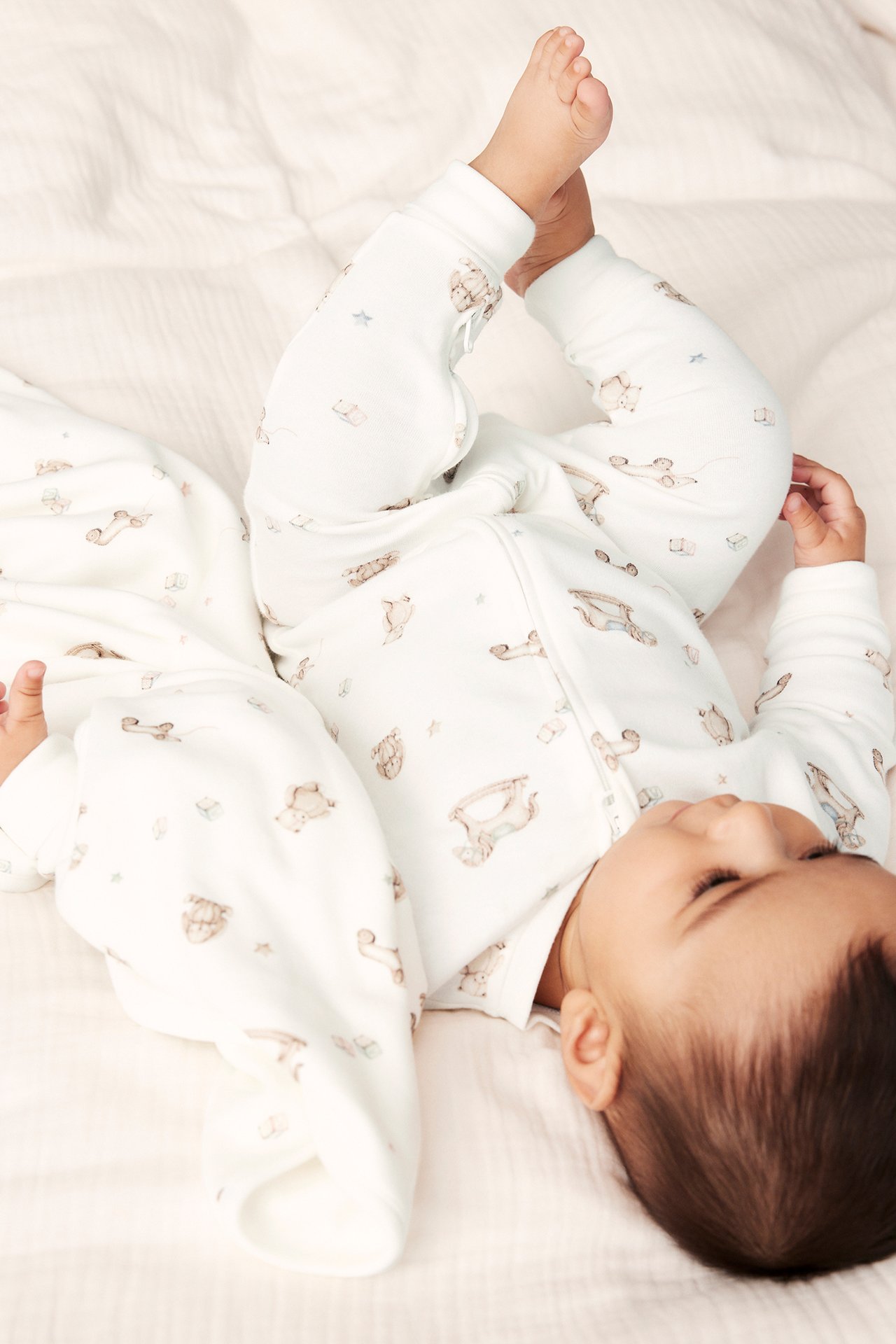 Top-down view of a baby in an off-white cotton romper with a toy pattern and front two-way zip.