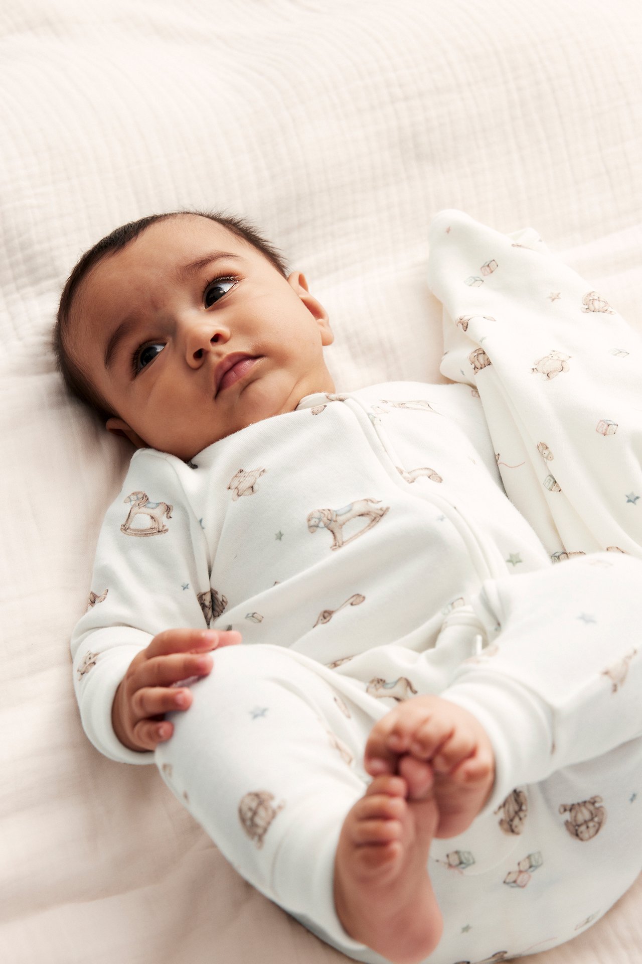 Front view of a baby in an off-white cotton pyjamas with a toy pattern and two-way zip.