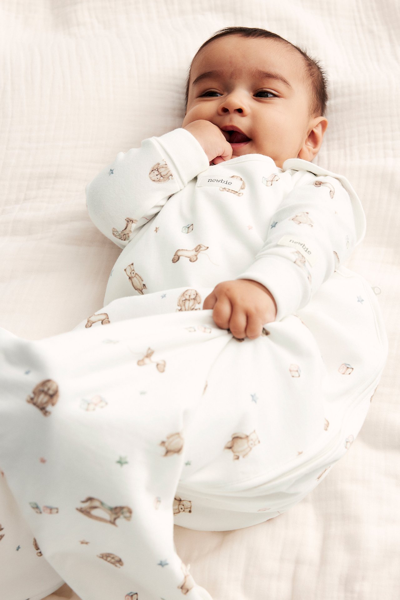 Vorderansicht eines Babys in einem cremefarbenen Bio-Baumwoll-Schlafsack mit niedlichem Spielzeugmuster.