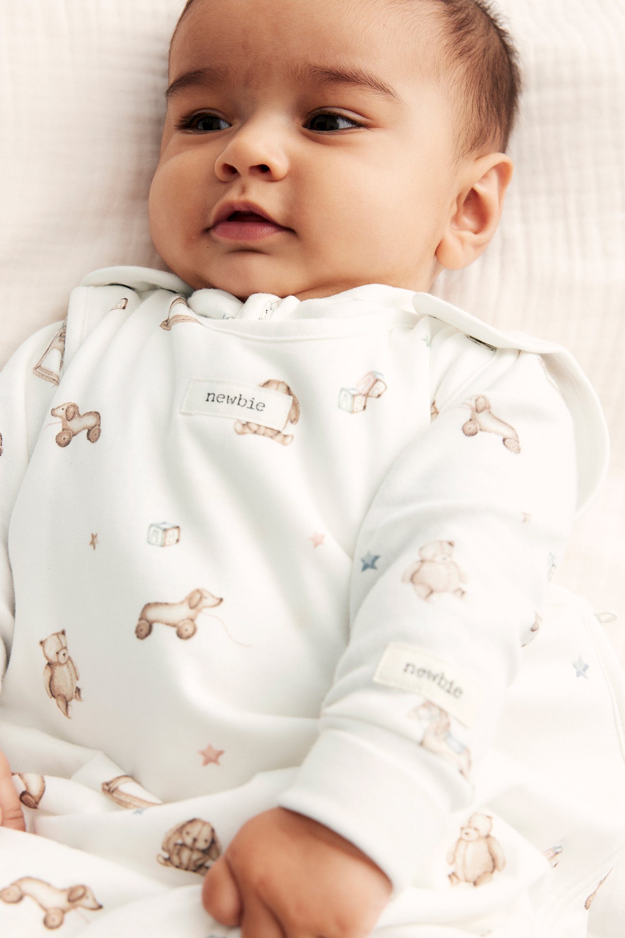 Nahaufnahme eines Babys in einem cremeweißen Schlafsack mit Spielzeugmuster und Schulter-Druckknöpfen.