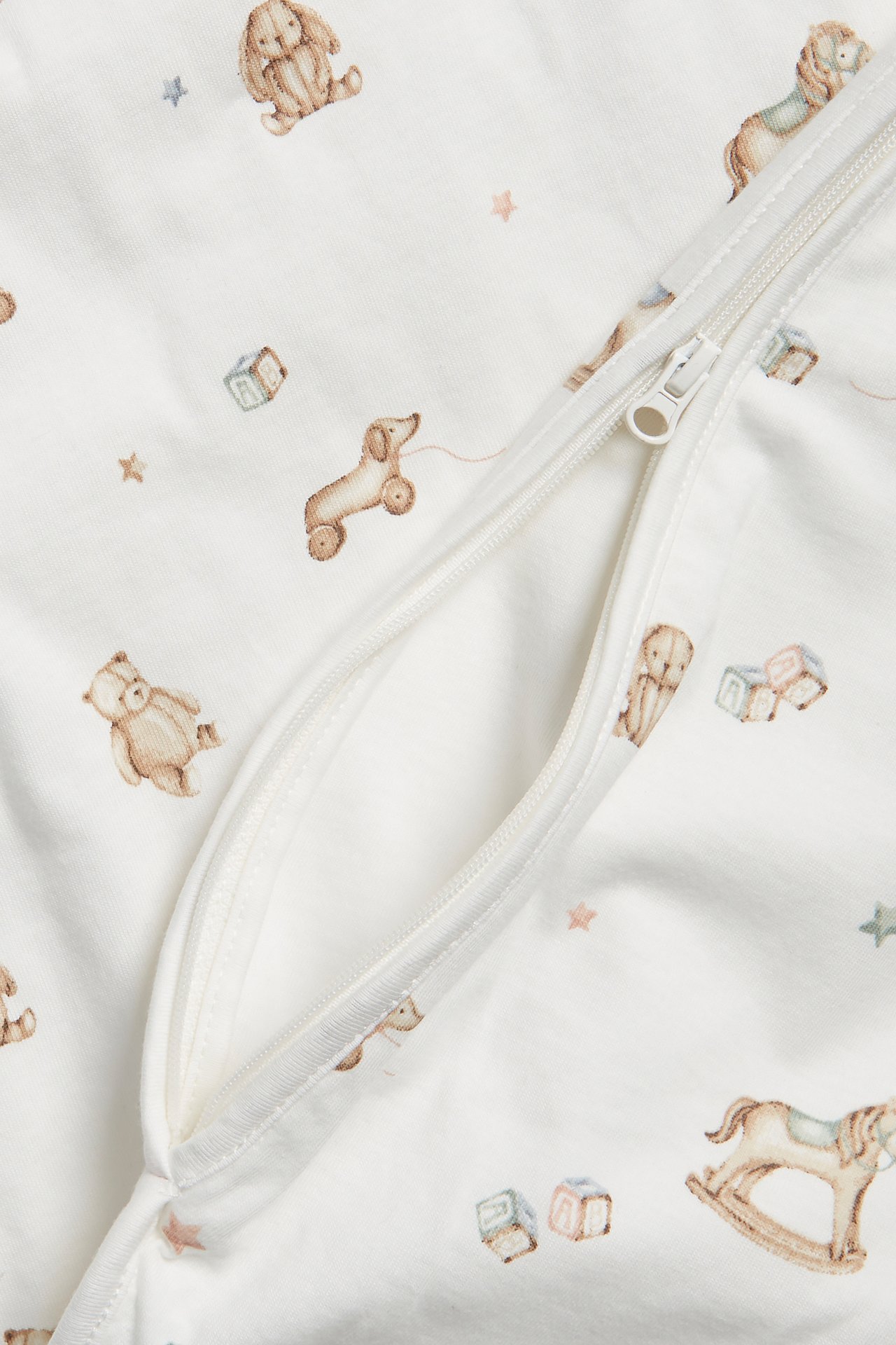 Nahaufnahme eines cremefarbenen Babyschlafsacks mit Spielzeugmuster und einem teilweise geöffneten weißen Reißverschluss.