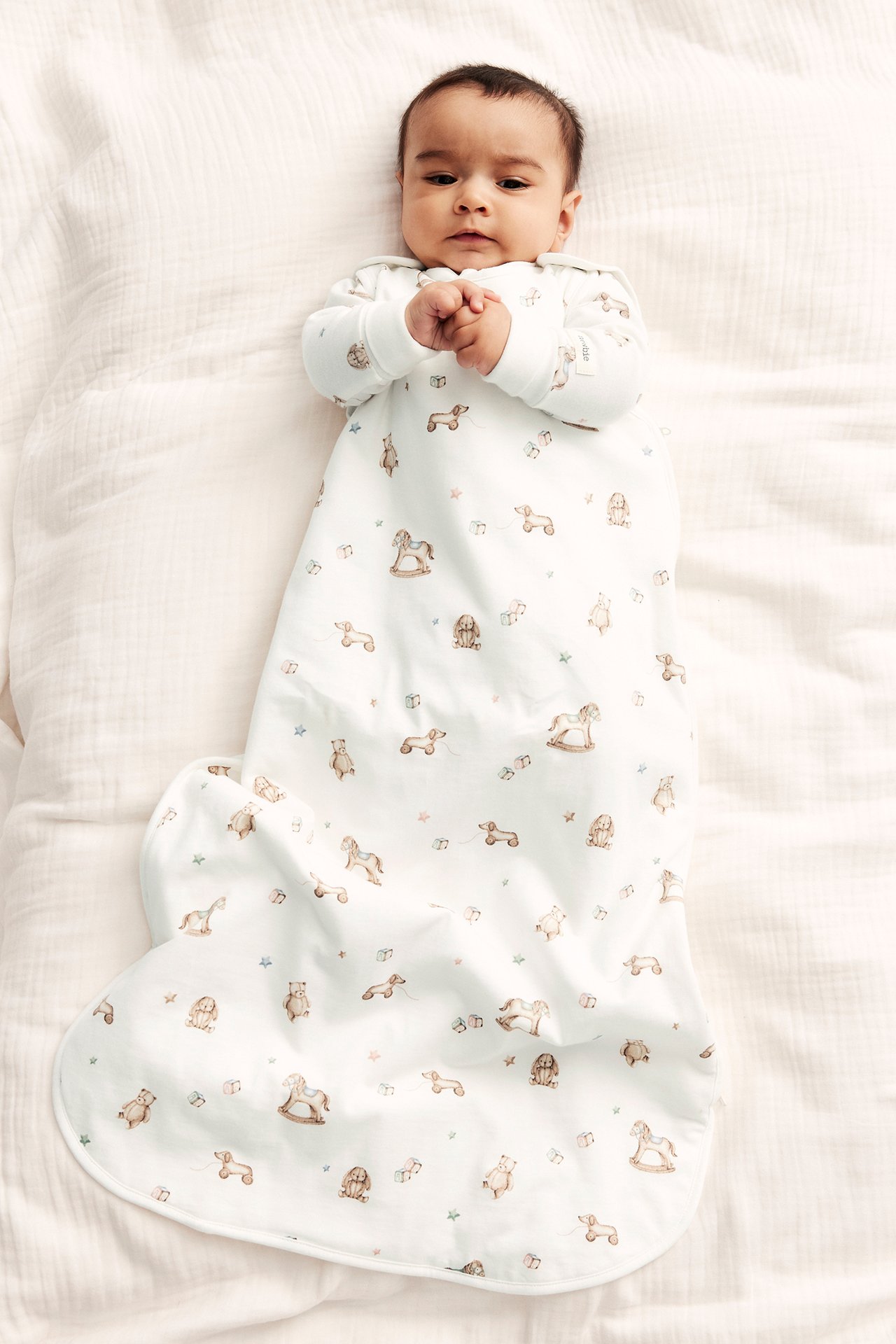 Draufsicht auf ein Baby in einem cremeweißen langärmeligen Schlafsack mit niedlichem Spielzeugmuster und Druckknöpfen.