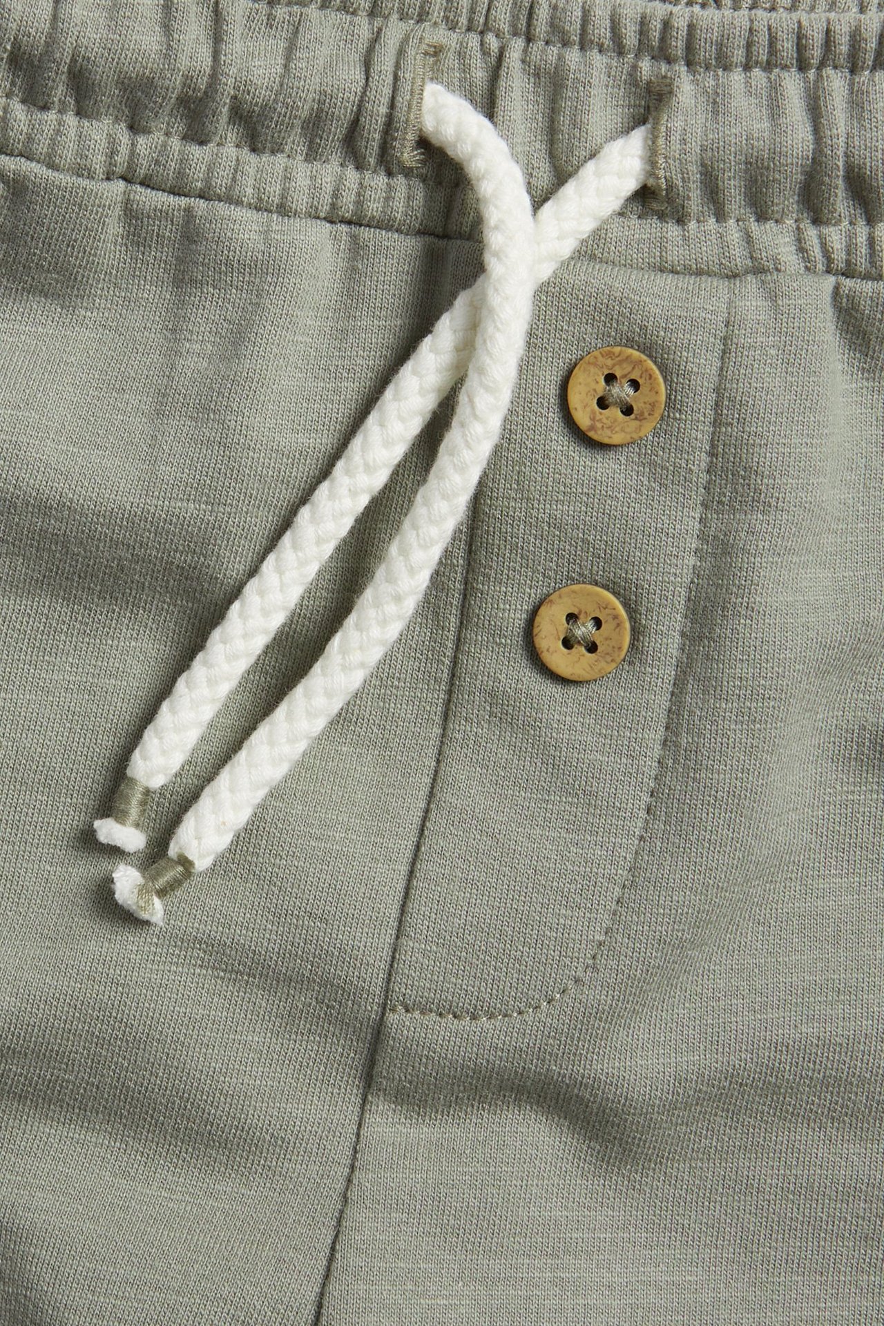 Close-up front view: green cotton shorts with elastic waist, white drawstring, and decorative wooden buttons.