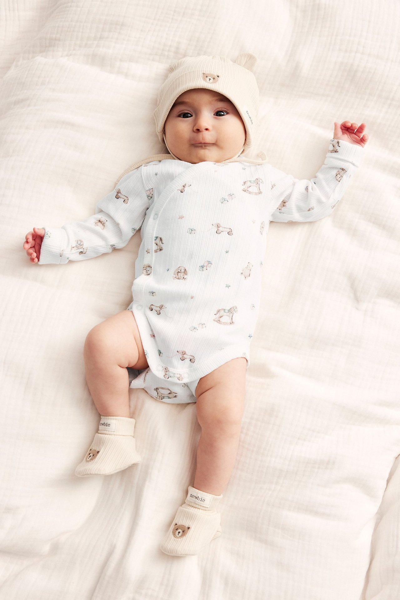 Front view of a baby wearing an off-white patterned wrap-around bodysuit, a beige bear-ear hat, and matching booties.