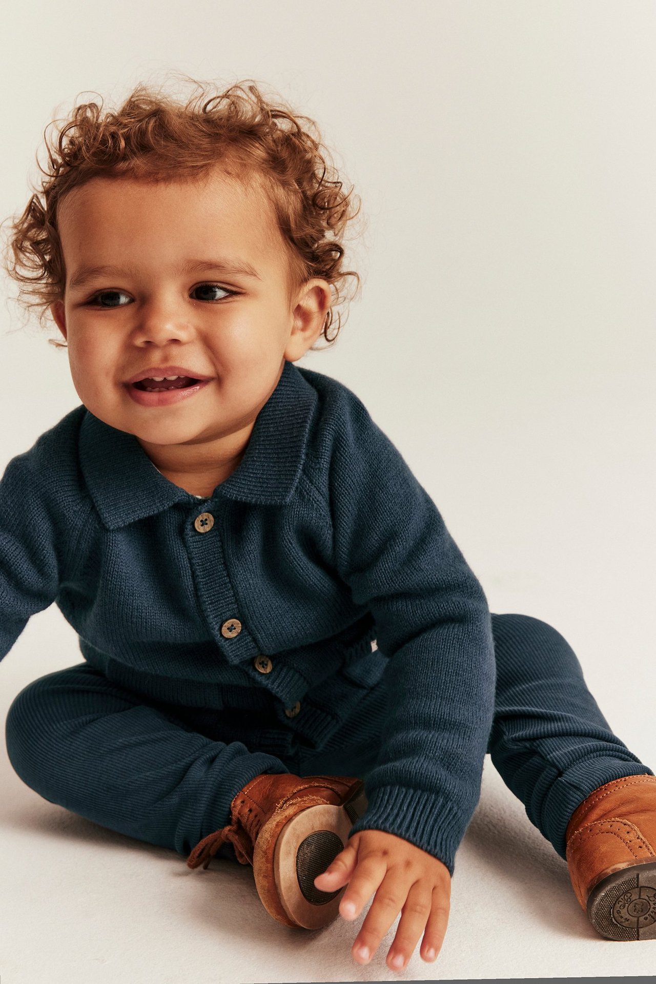 Front view of a smiling baby in a dark blue knitted cardigan with buttons, matching trousers, and brown shoes.