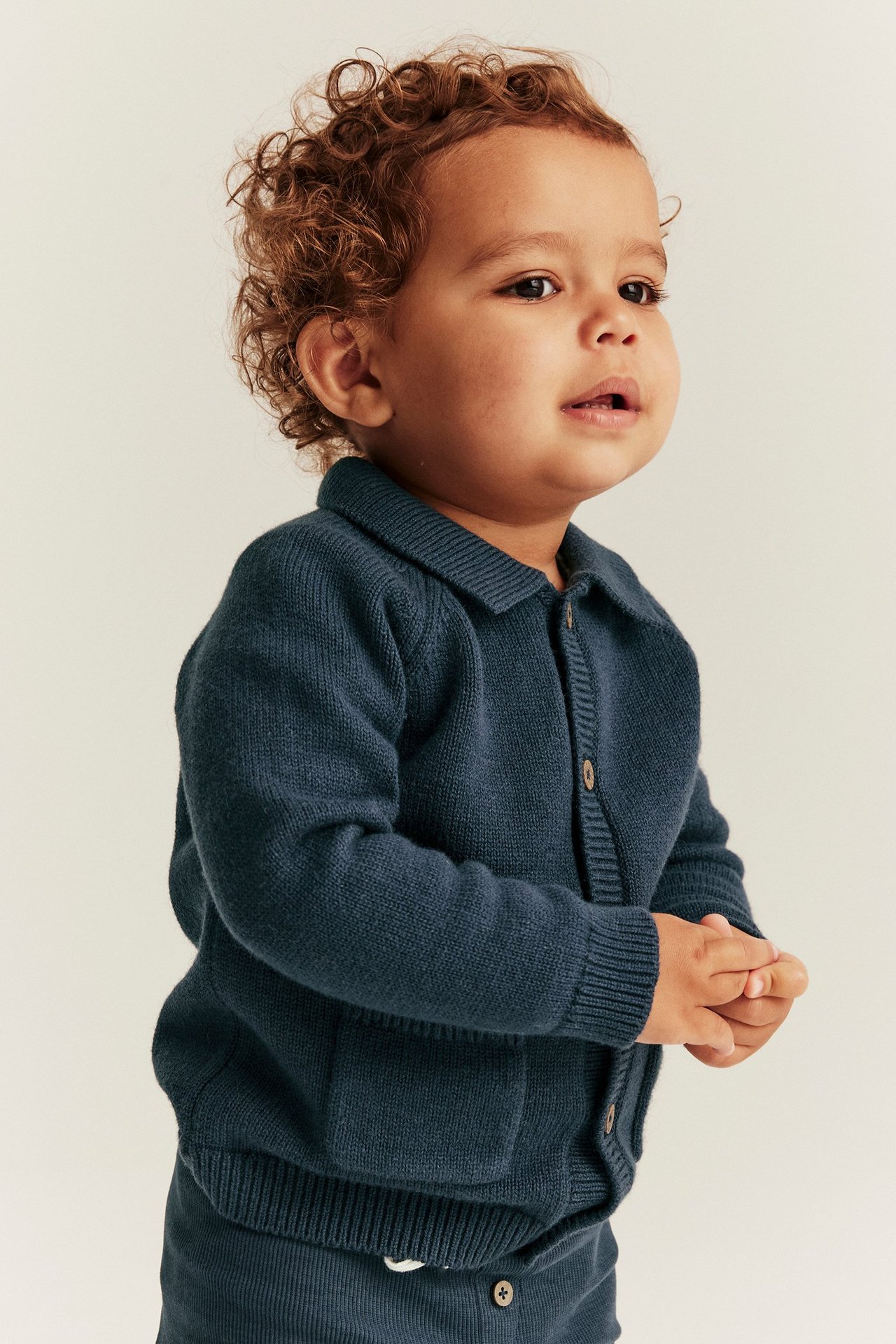 Side view of a baby in a dark blue knitted cardigan with collar, buttons, and pockets, over matching trousers.