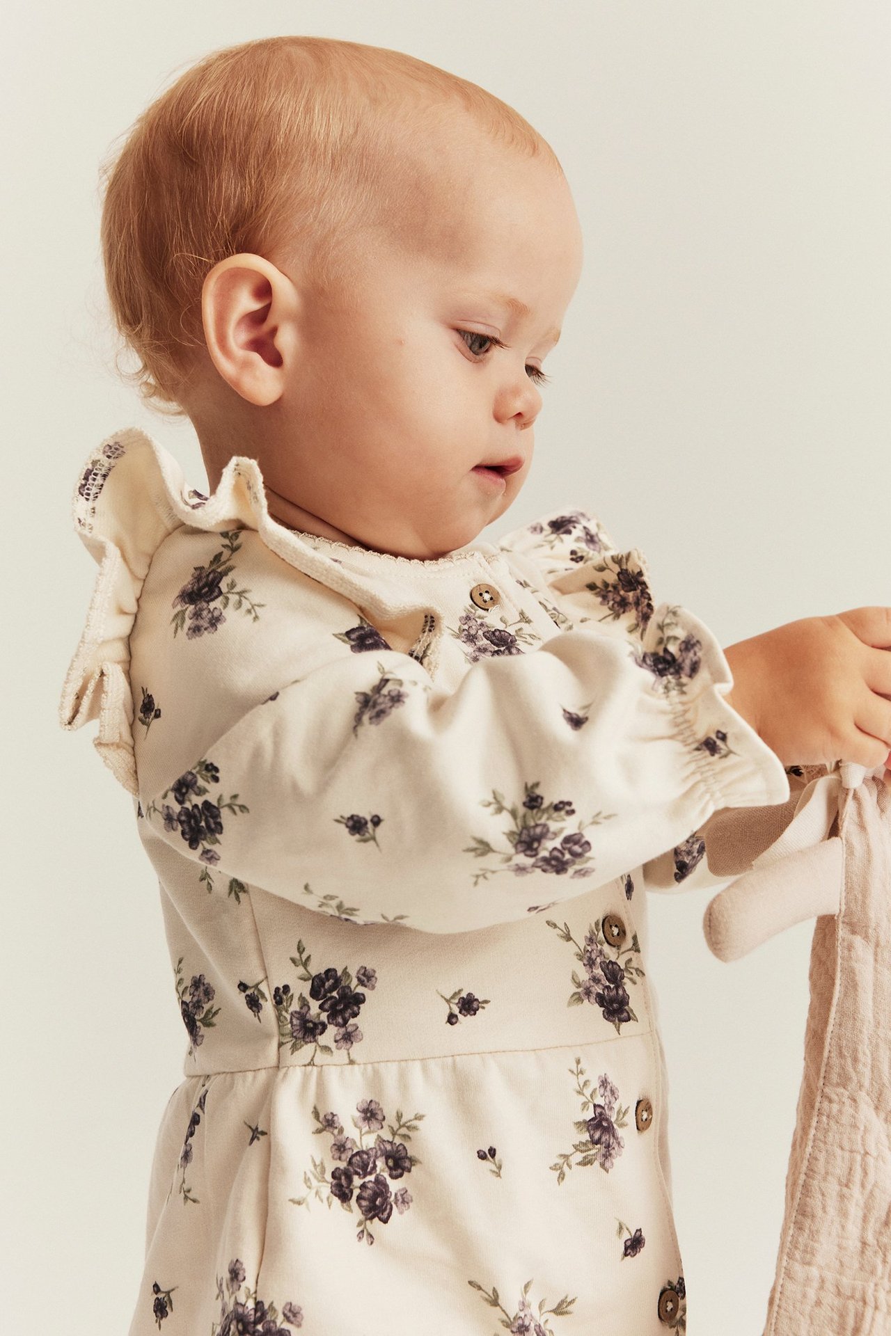 Seitenansicht eines Babys in hellbeigem geblümtem Baumwoll-Overall mit Rüschen, hält ein rosa Stofftier.