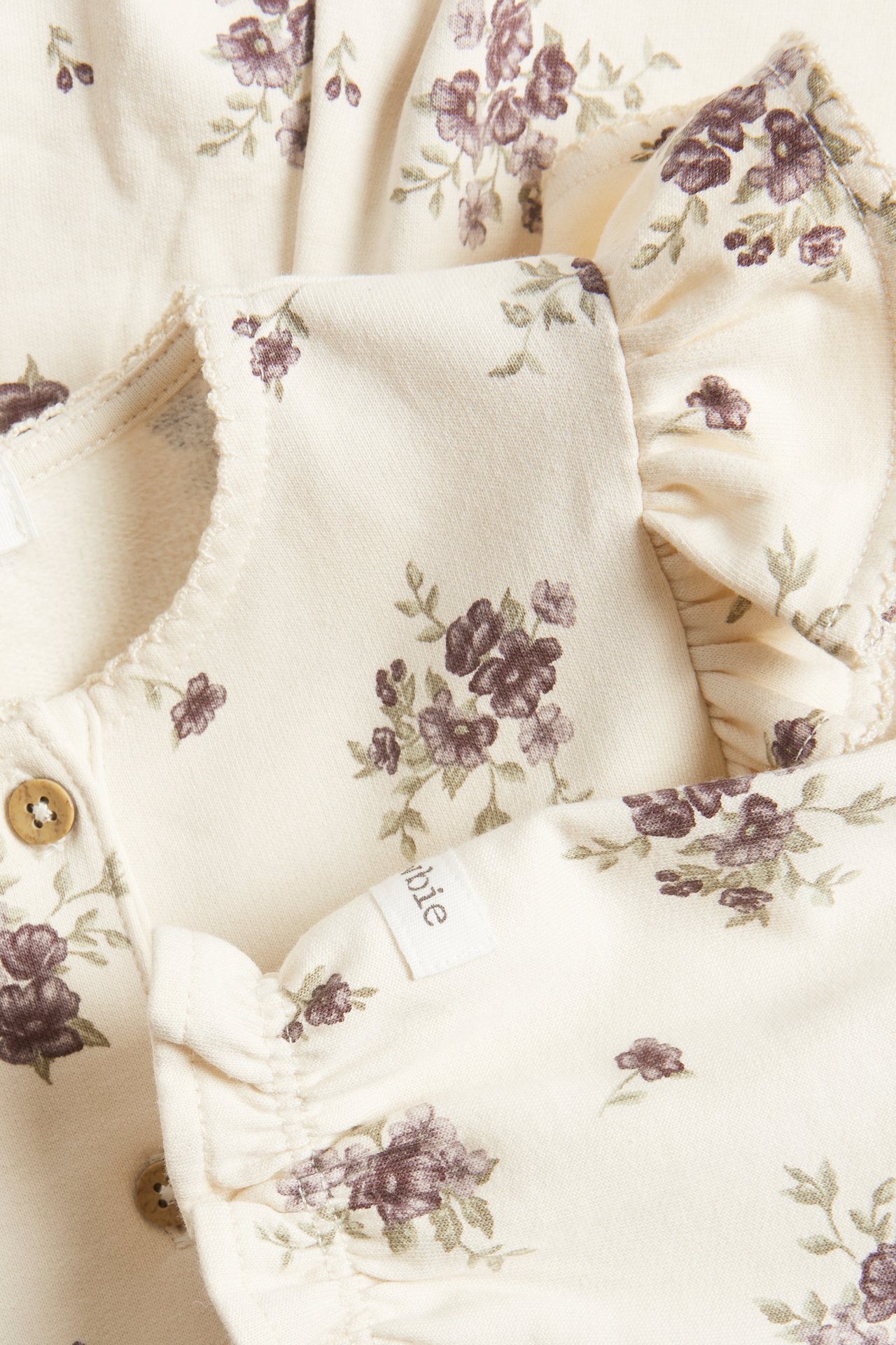 Close-up of a light beige baby floral cotton jersey jumpsuit with ruffles and front buttons.