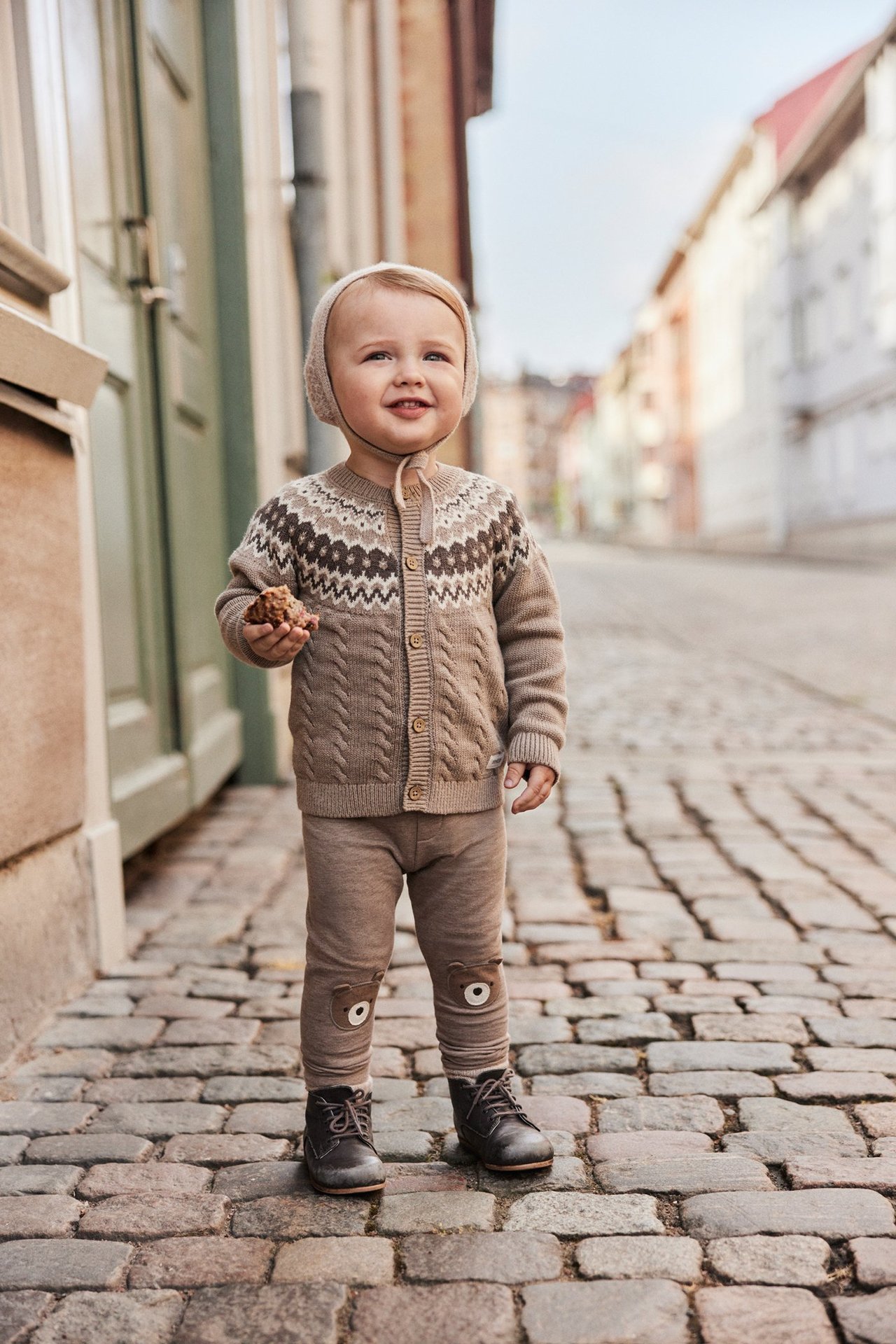 Vorderansicht eines Babys in brauner Jacquard-Strickjacke, beigen Leggings, dunklen Stiefeln und Mütze, hält Gebäck.