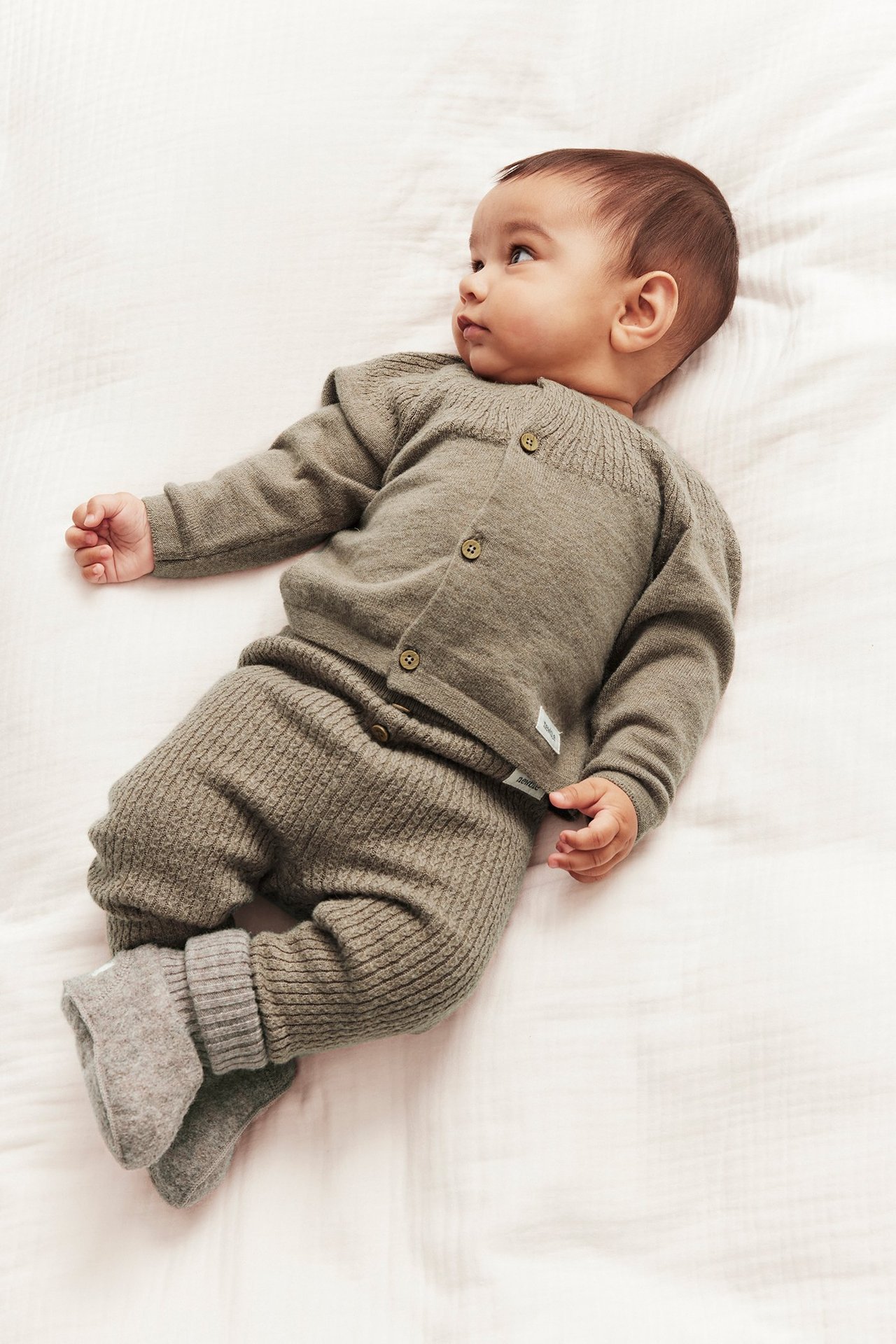 Seitenansicht eines Babys in einer melierten braunen Strickjacke mit Knöpfen und passender Strickhose und Schühchen.