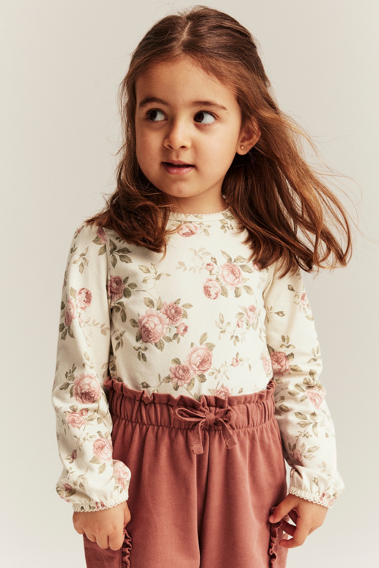 Front view of a kid in a light beige floral long-sleeved top with puff sleeves and dusty pink trousers.
