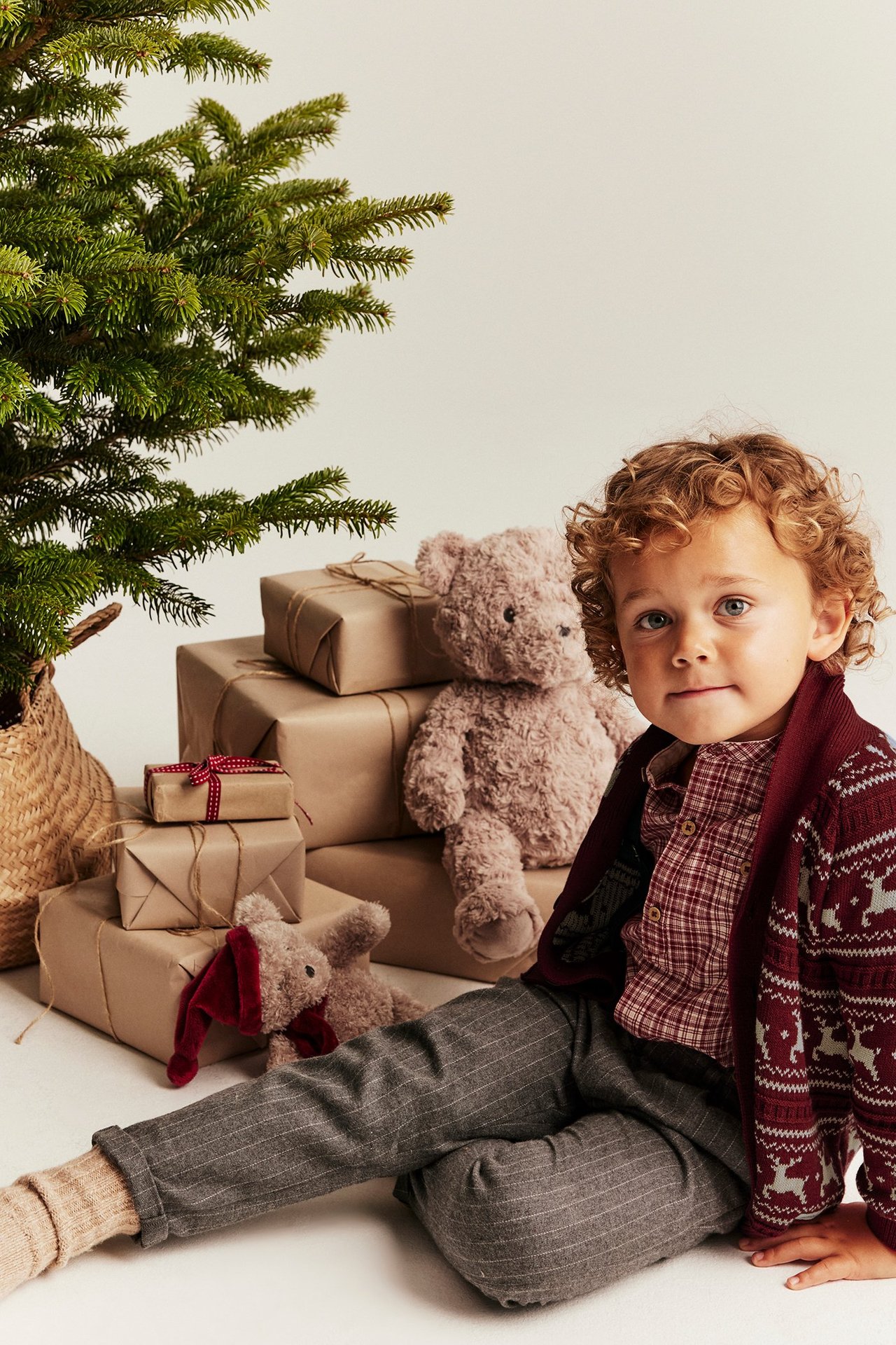 Kid wears red jacquard knit reindeer cardigan, plaid shirt, grey pinstripe pants, beige socks.