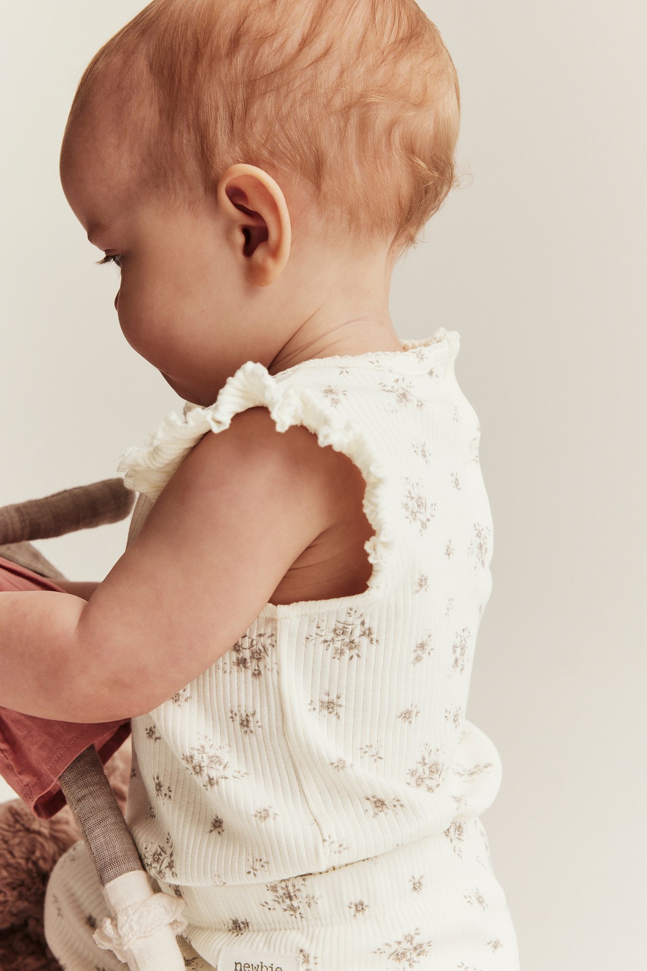 Side view of a baby in an off-white ribbed floral bodysuit with ruffle sleeves, matching leggings, holding a doll.