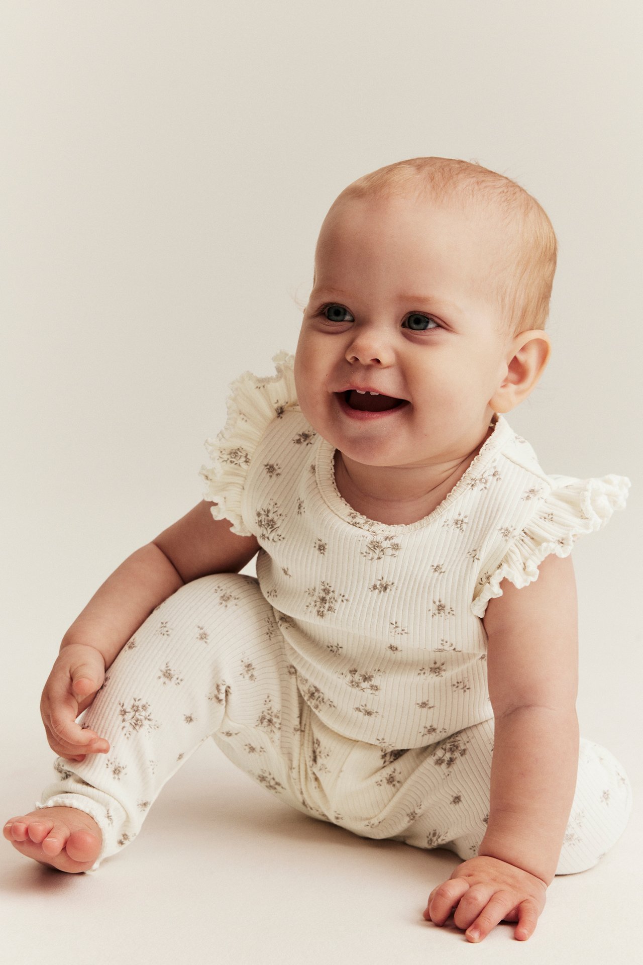 Front view of a smiling baby in an off-white ribbed floral bodysuit with ruffle sleeves and matching leggings.