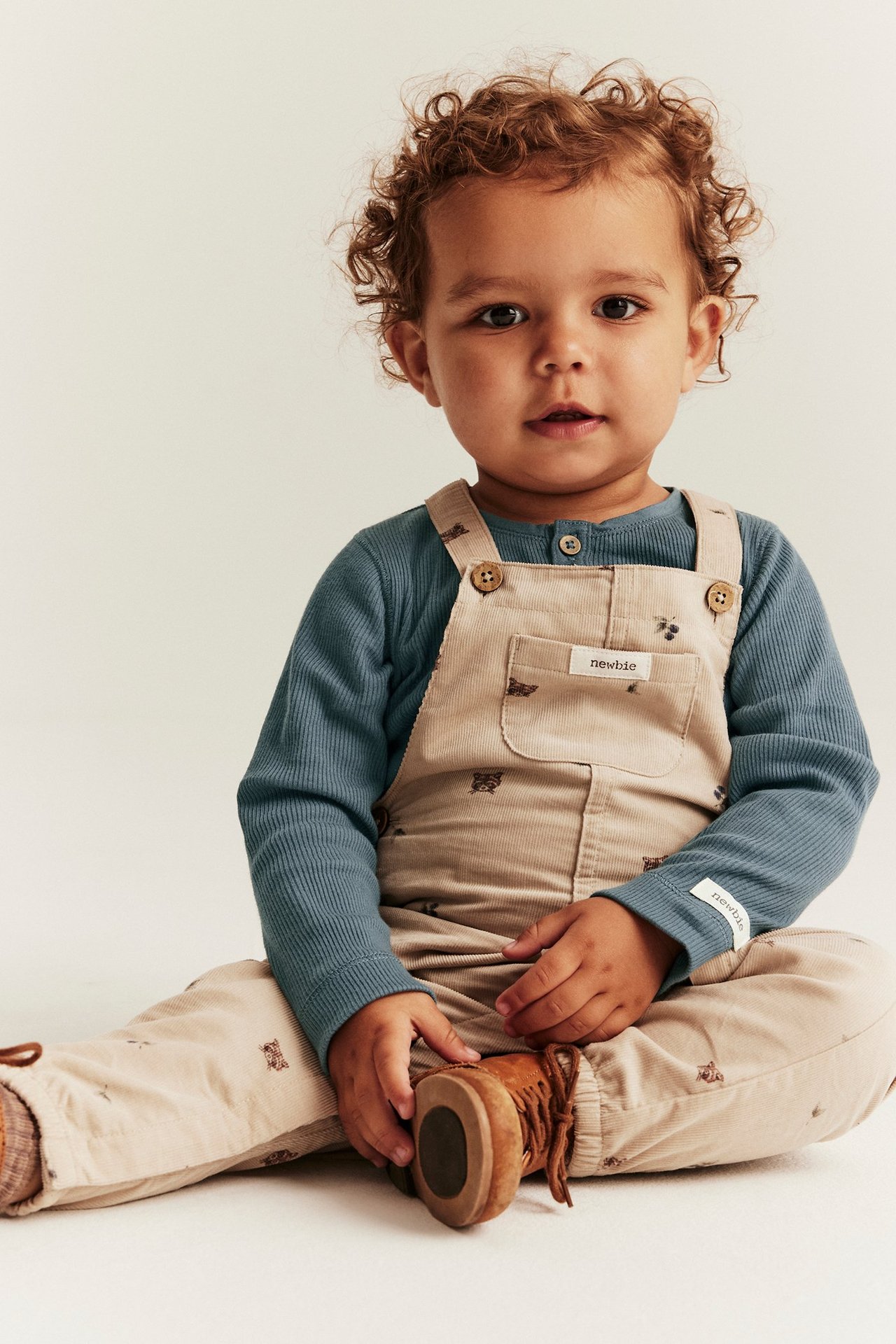 Front view of a baby in a blue ribbed long-sleeved body, beige corduroy overalls, and brown shoes.