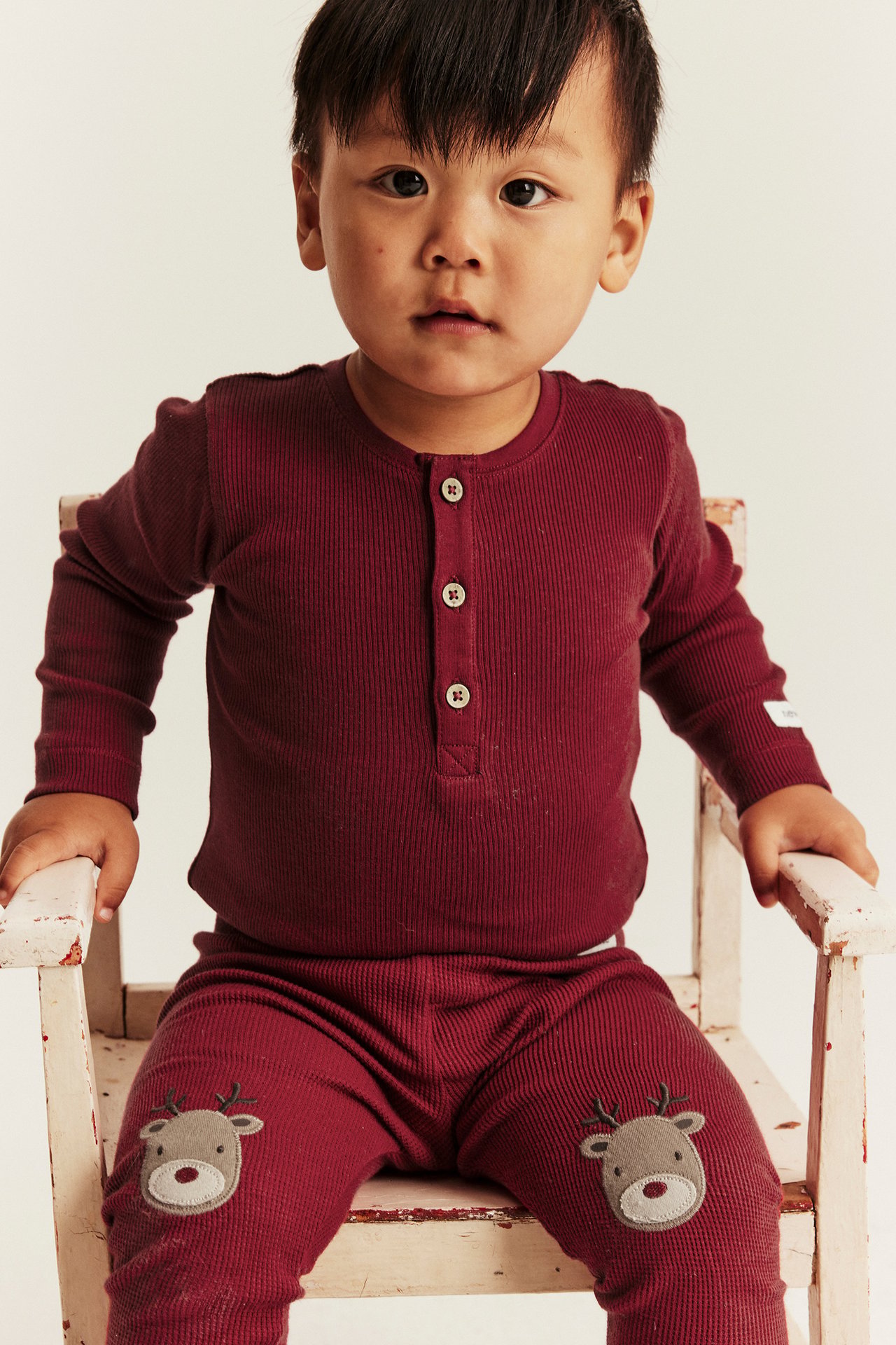 Vorderansicht eines Babys in weinrotem, geripptem Langarmshirt und passender Hose mit Rentierknien.