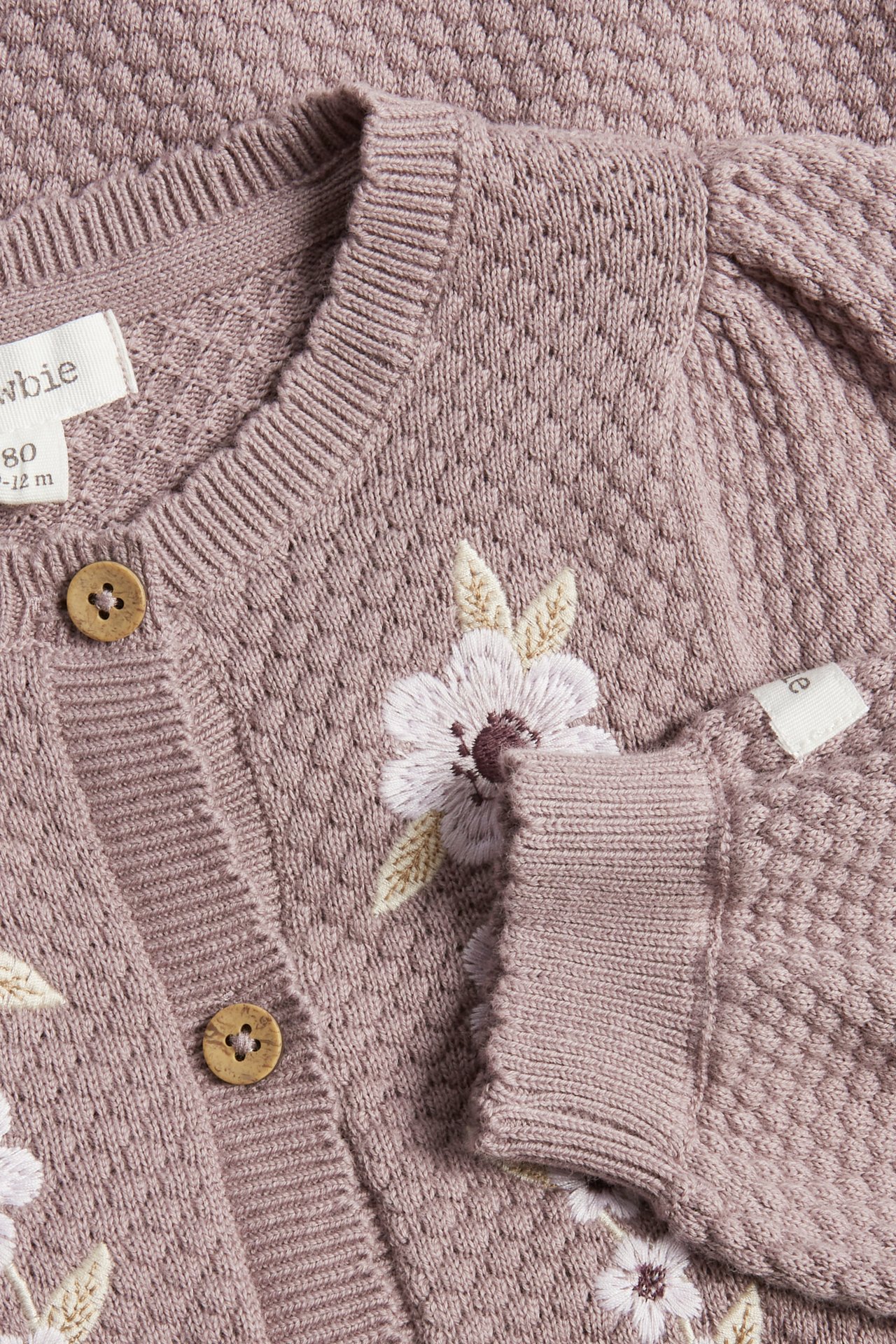 Nahaufnahme eines helllila strukturstrick Baby-Overalls mit Blumenstickerei und Holzknöpfen vorne.
