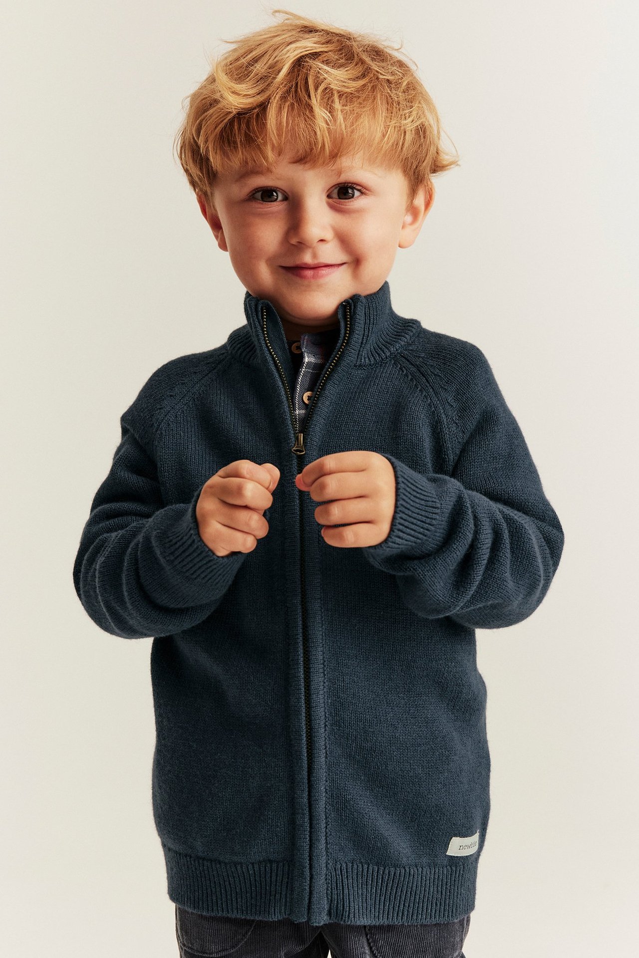 Vorderansicht: Kind in dunkelblauer Strickjacke aus Wollmix mit Reißverschluss über kariertem Hemd, hält Reißverschluss.