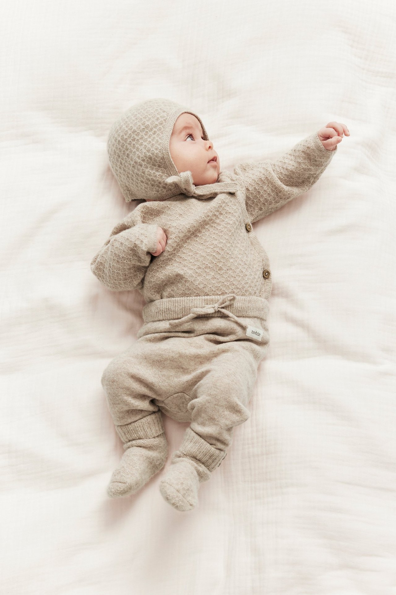 Baby wears beige melange wool-cashmere knit drawstring pants, cardigan, bonnet, and booties.